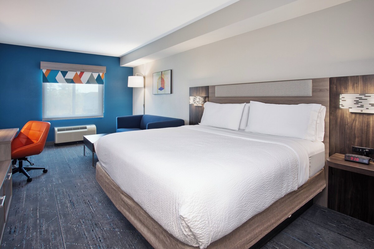 A hotel room with a large bed, a desk, an orange chair, a window with a geometric curtain, and a wall-mounted lamp.