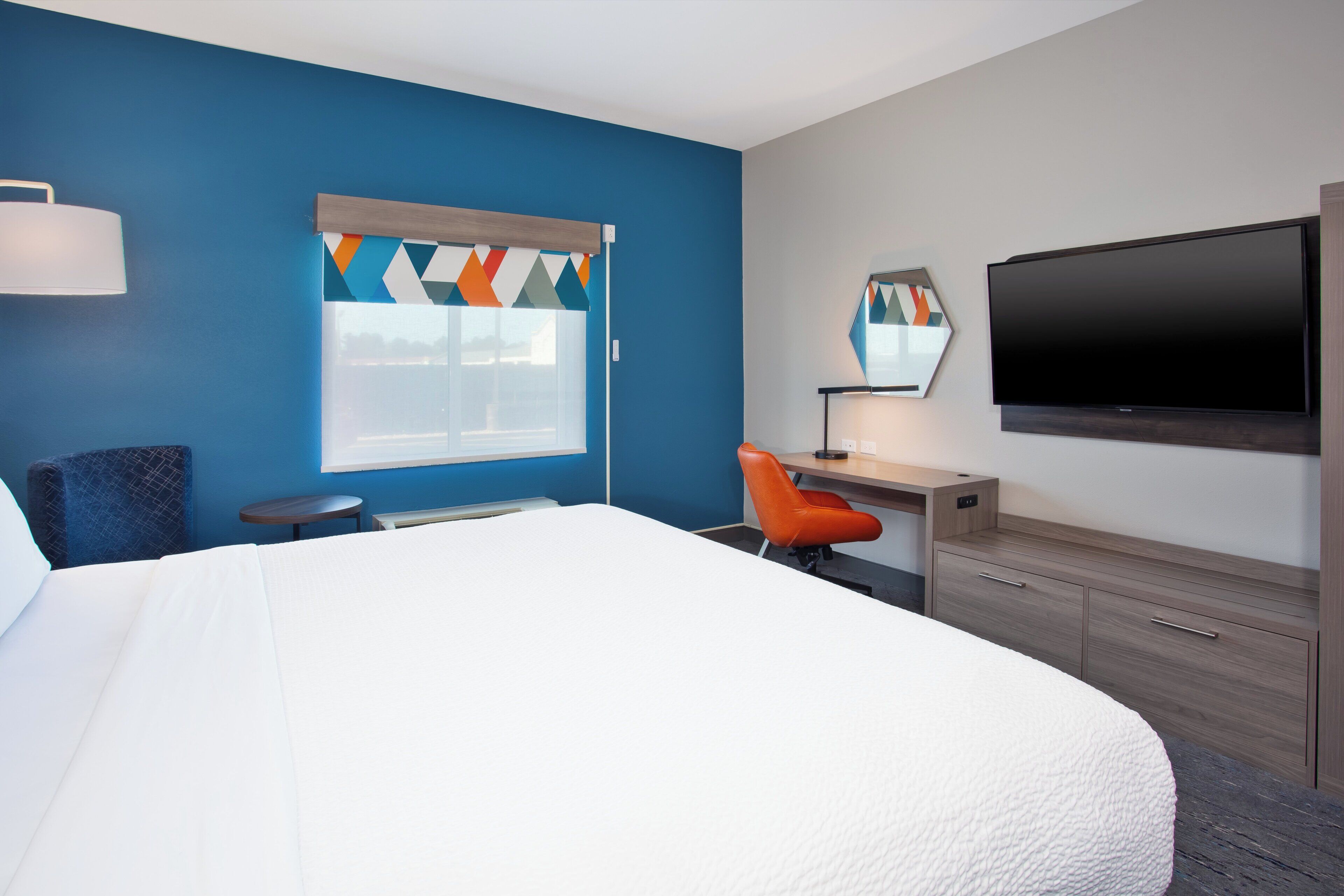 Habitación de hotel con una cama grande, un escritorio con silla, un televisor de pantalla plana y una ventana con cortina geométrica.