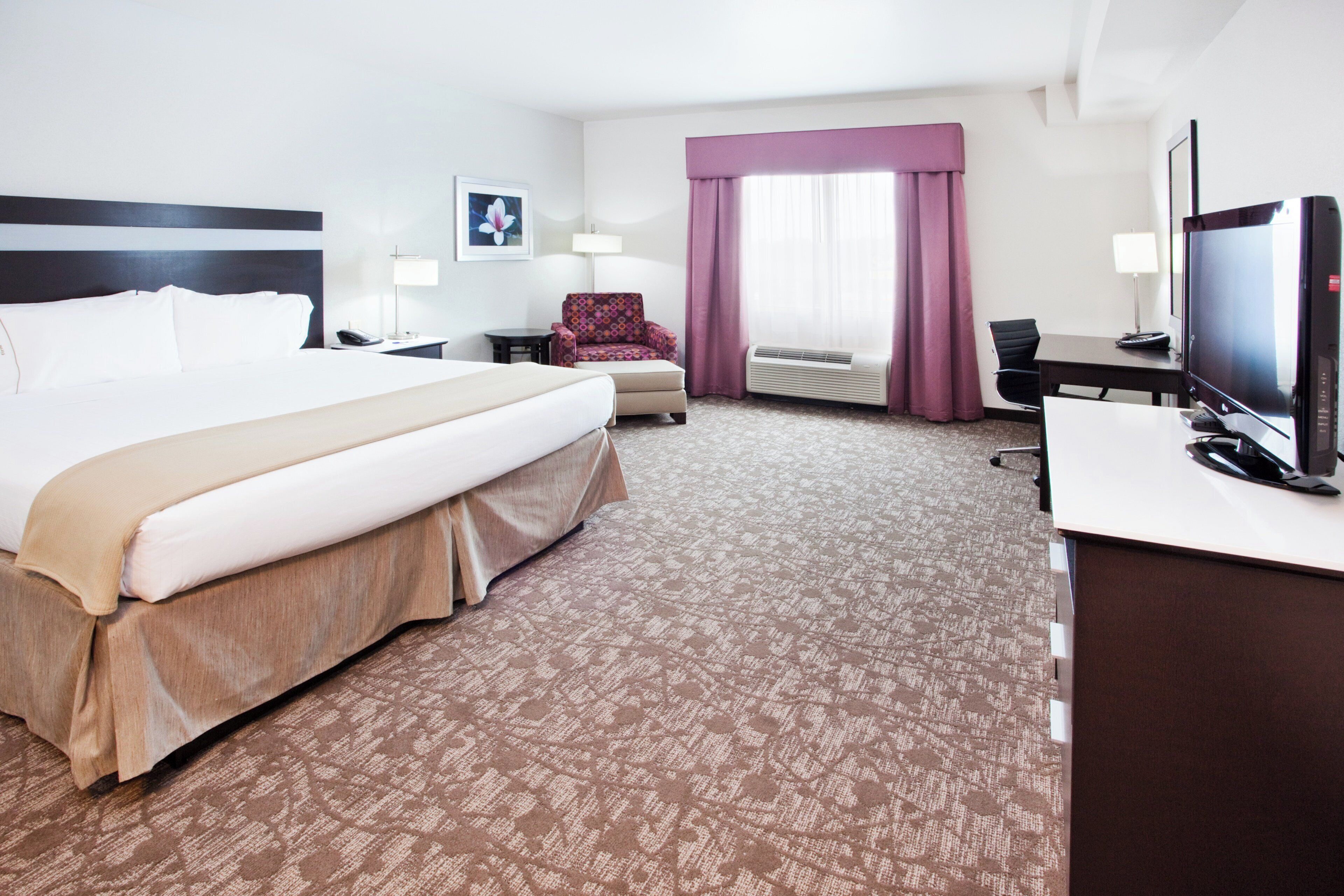 Habitación de hotel con una cama grande, un escritorio con computadora, una silla, una lámpara de escritorio, una ventana con cortinas y un televisor montado en la pared.