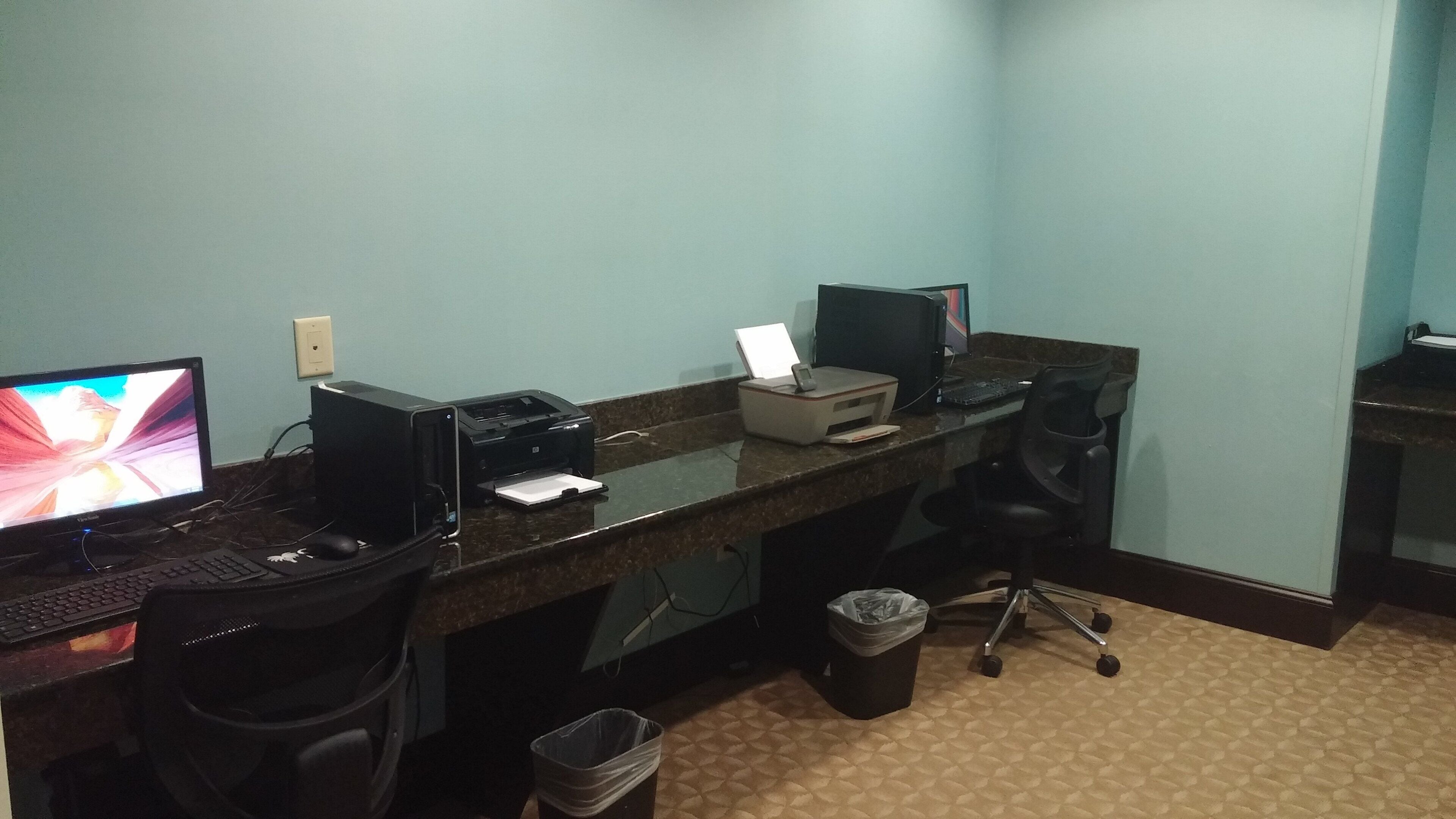 A hotel room with a desk, two monitors, a printer, a trash bin, and a chair.