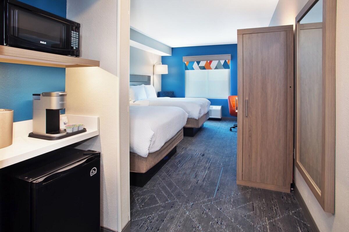 A compact hotel room with a kitchenette, a bed, a wardrobe, and a window with a colorful curtain.
