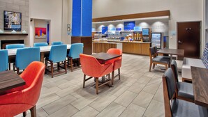A modern hotel dining area with blue and orange chairs, a central wooden table, and a breakfast bar with various food items. - Holiday Inn Express Augusta North by IHG (Augusta)