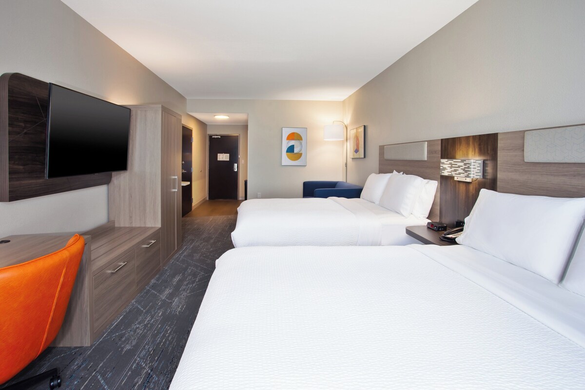 A hotel room with two beds, a desk, an orange chair, a flat-screen TV, and artwork on the walls.