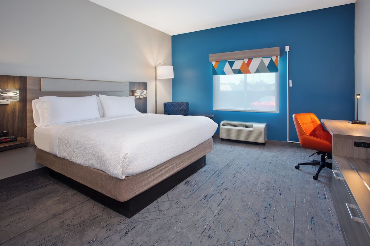 A modern hotel room with a large bed, a desk with a chair, and a window with a geometric curtain.