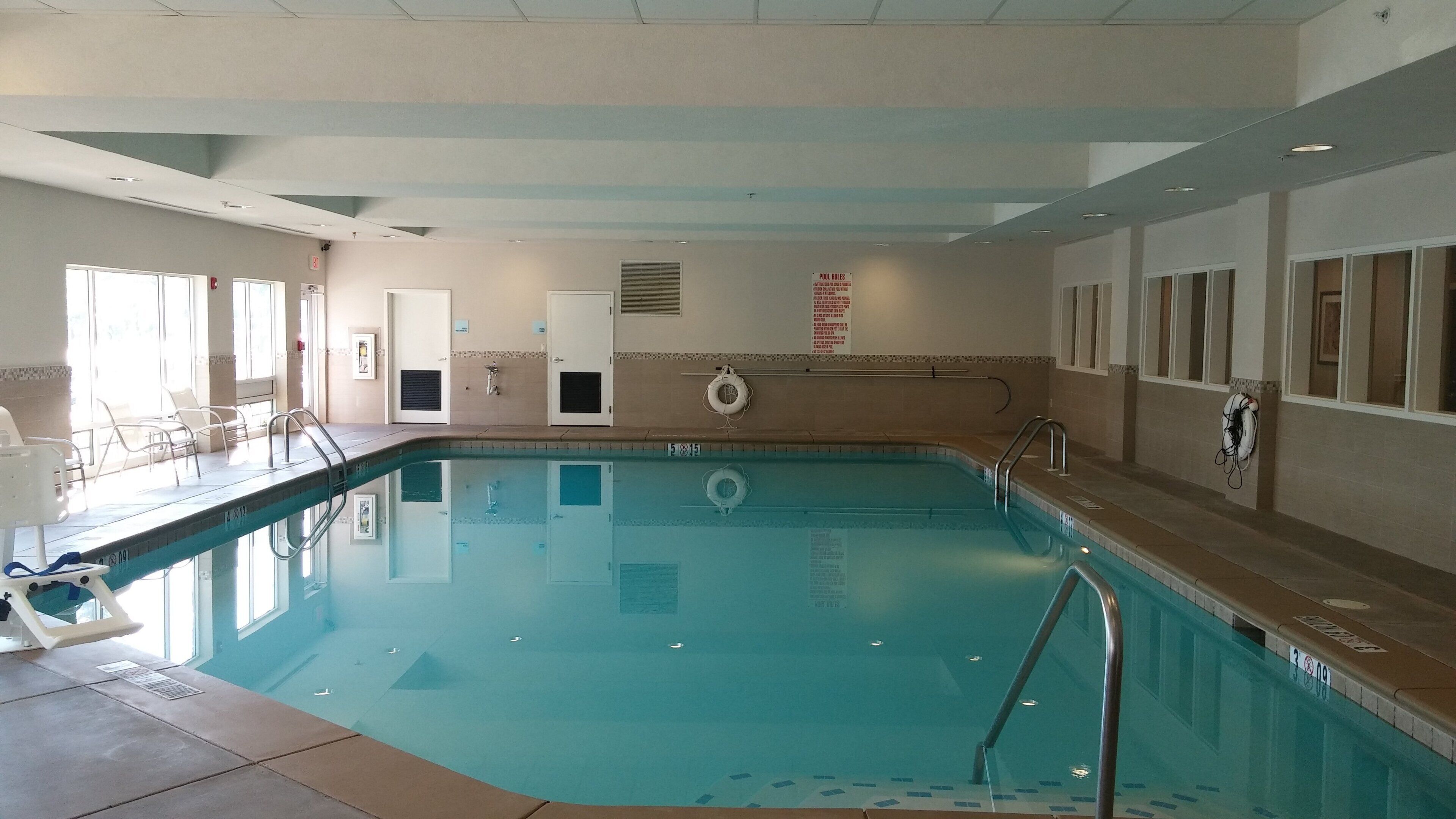 A swimming pool with clear water, surrounded by a tiled deck and a row of chairs.