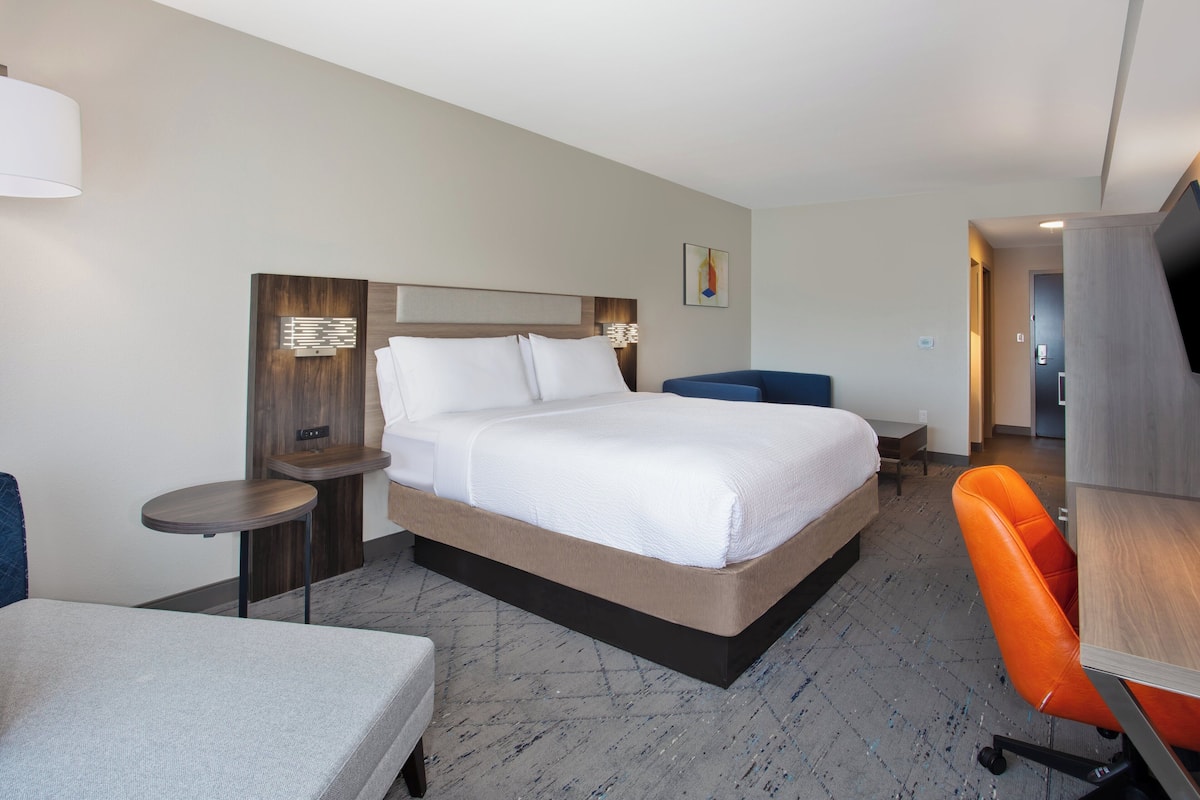 A hotel room with a large bed, a desk, an orange chair, and a television.