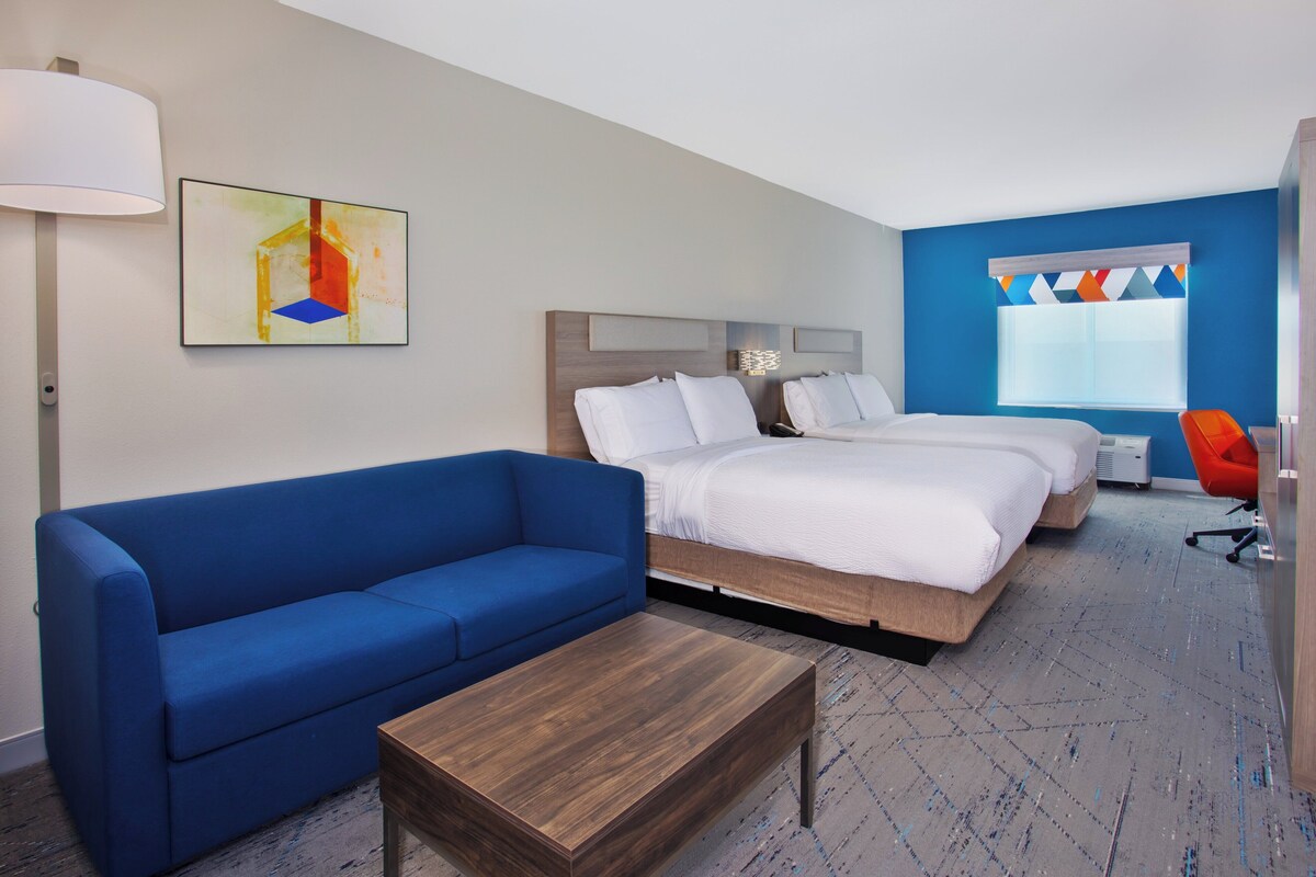 A hotel room with a blue sofa, two beds, a wooden coffee table, and a colorful abstract painting.