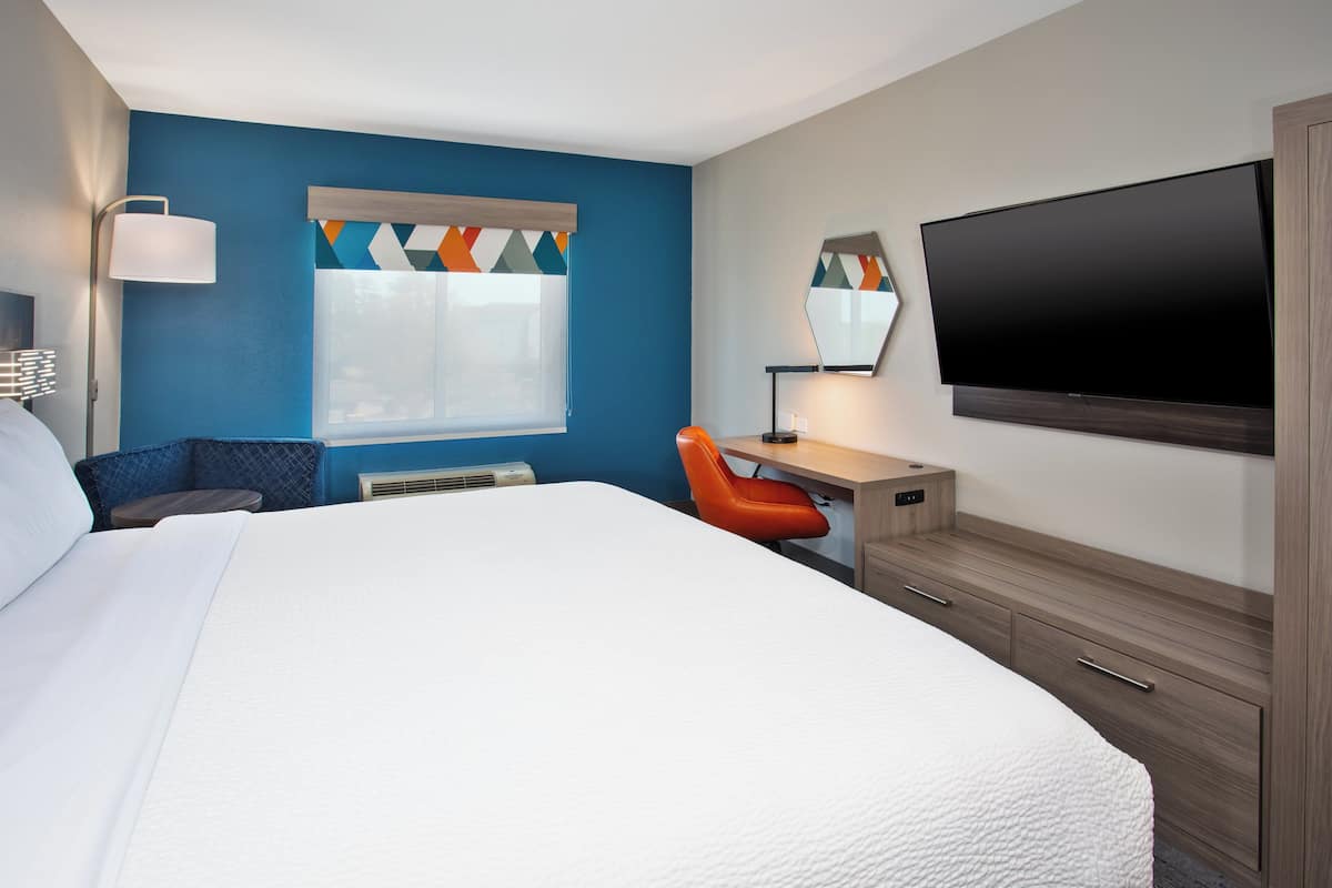 A hotel room with a large bed, a desk with a chair, a flat-screen TV, a window with blinds, and a mirror on the wall.