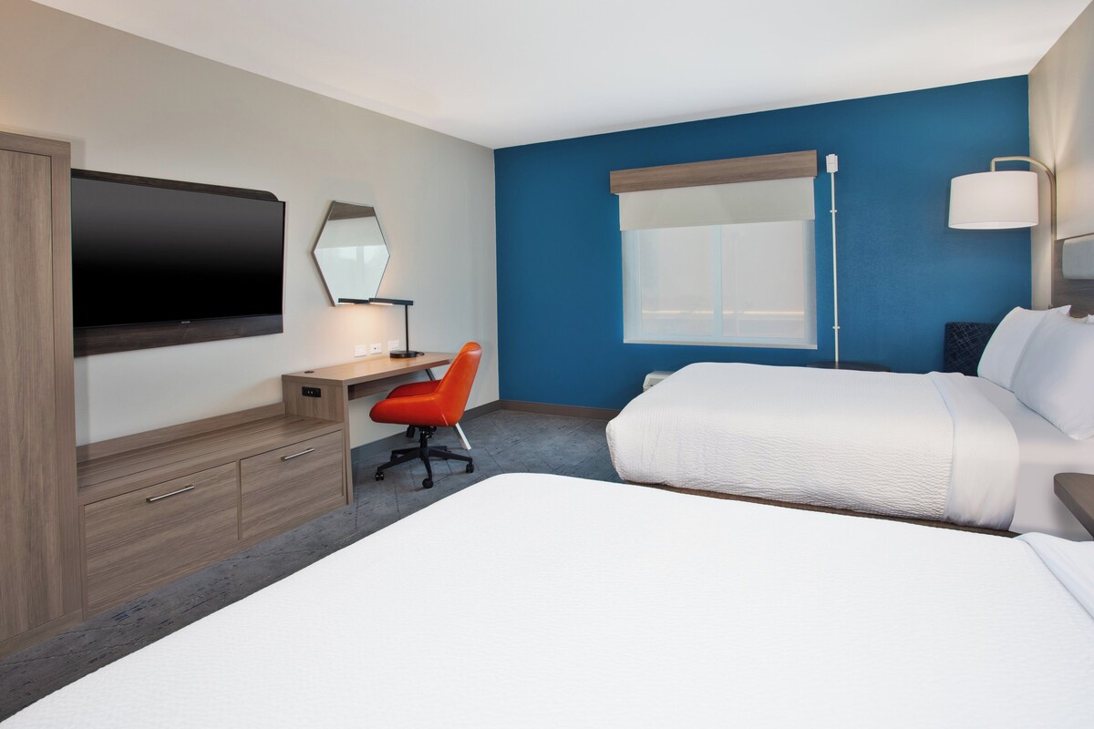 A hotel room with a large bed, a desk with a chair, a flat-screen TV, a bedside table, and a window with a view.