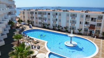 Una piscina con fuente, rodeada de sillas de descanso y palmeras, con vista al mar y edificios al fondo.