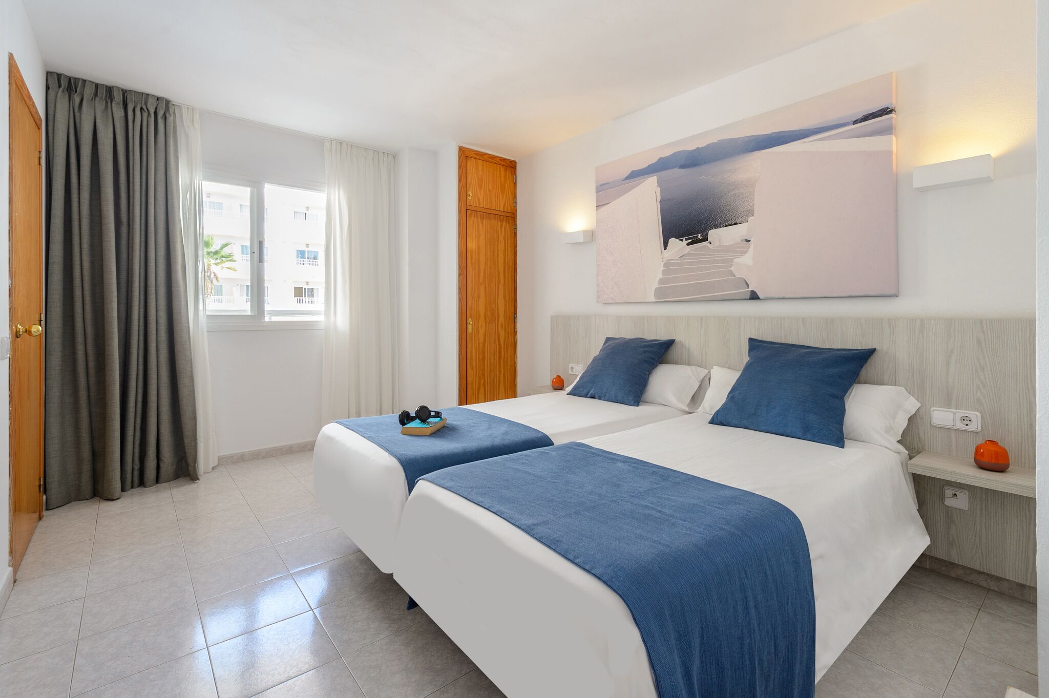 A hotel room with two beds, a large window with curtains, a painting on the wall, and a nightstand with a lamp.