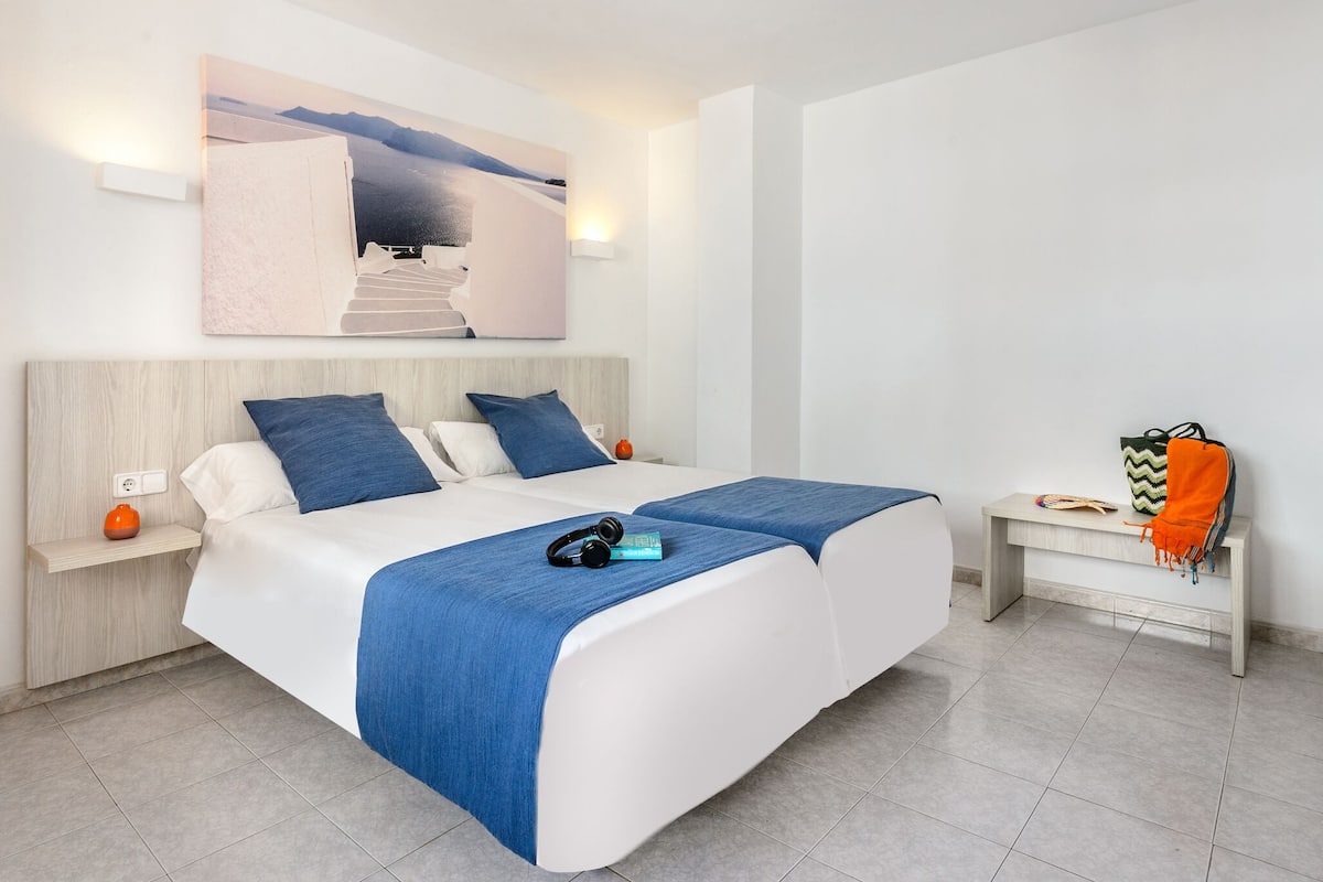 Una habitación de hotel moderna con una cama grande, un banco con una manta, una obra de arte en la pared y una mesita con una lámpara.