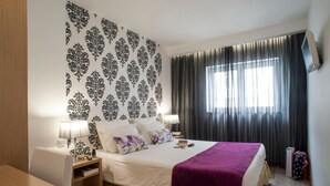 Habitación de hotel con cama, dos mesitas de noche, un escritorio, una silla, televisor montado en la pared y una ventana con cortinas.