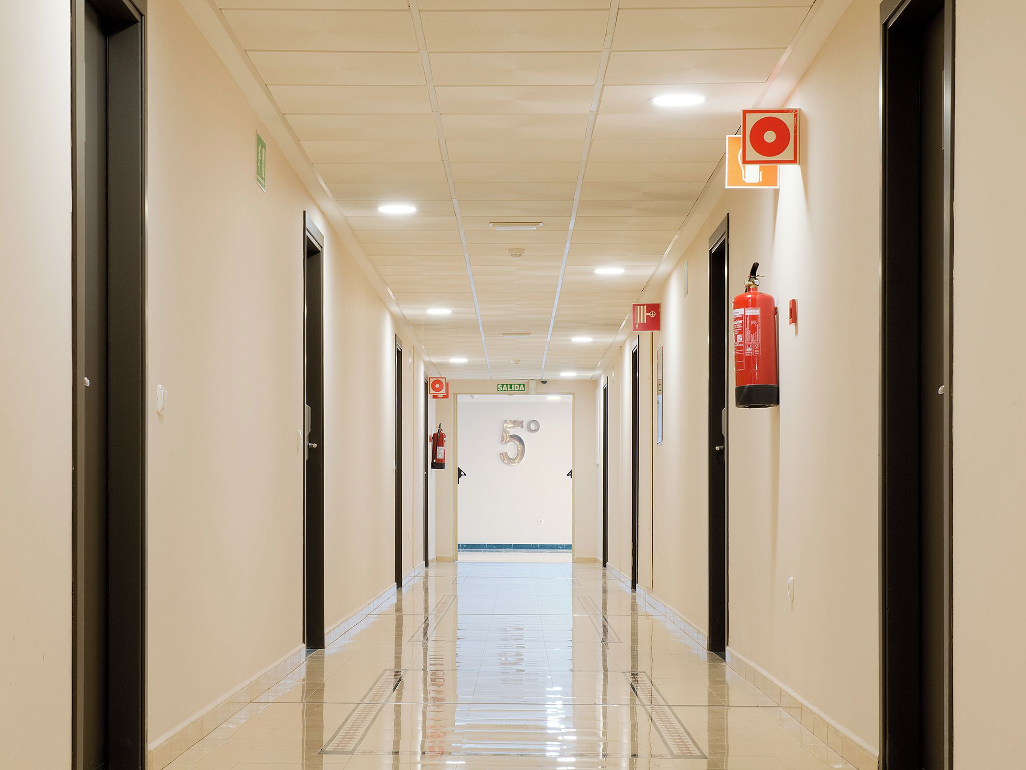 A clean, well-lit hallway with doors on both sides, a fire extinguisher mounted on the wall, and a sign indicating the direction to the exit.