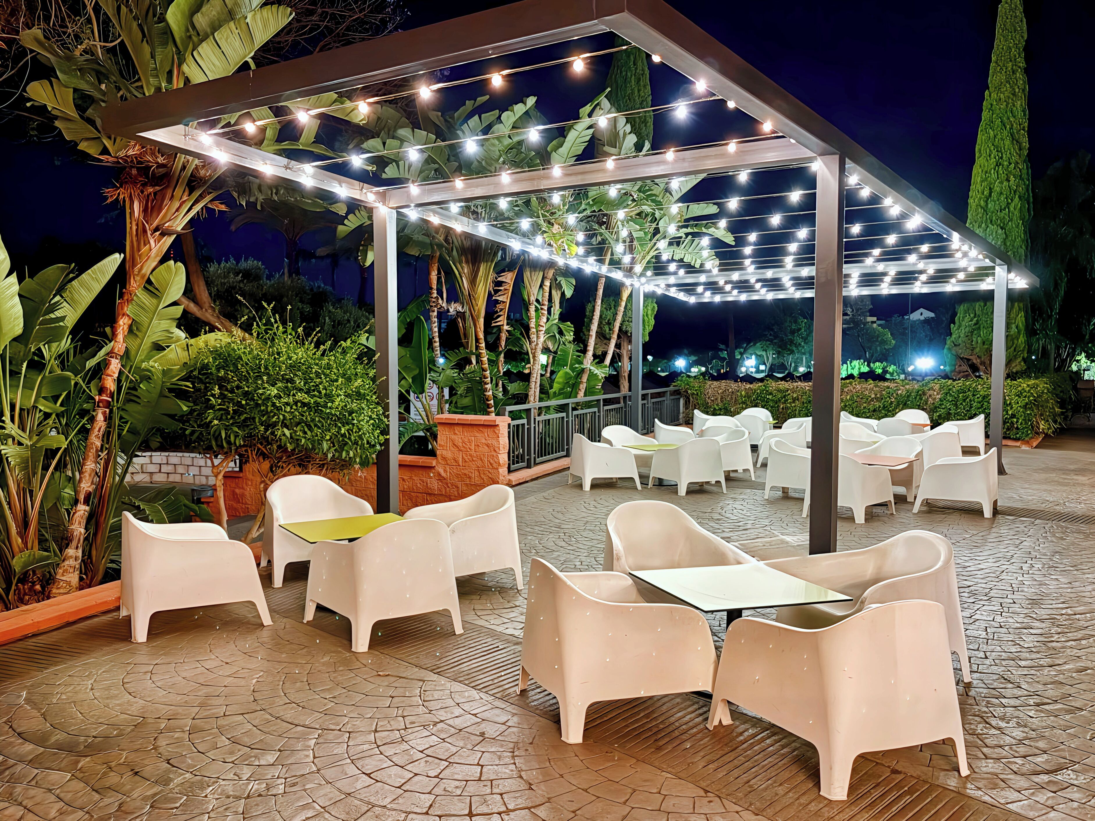 An outdoor patio with white plastic chairs and tables, string lights, and tropical plants.