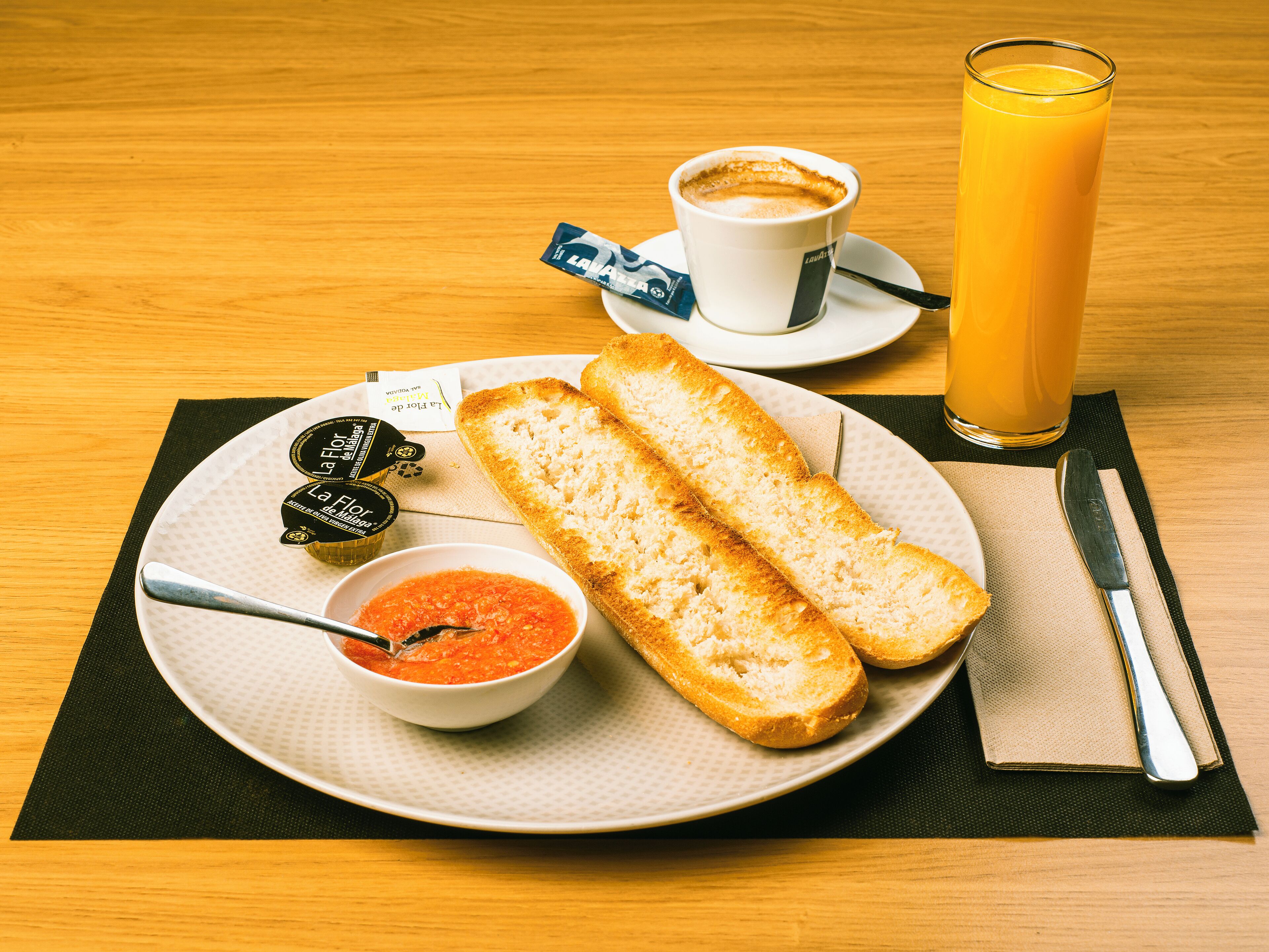 Un desayuno con tostadas, un tazón de sopa de tomate, una taza de café y un vaso de jugo de naranja sobre una mesa de madera.