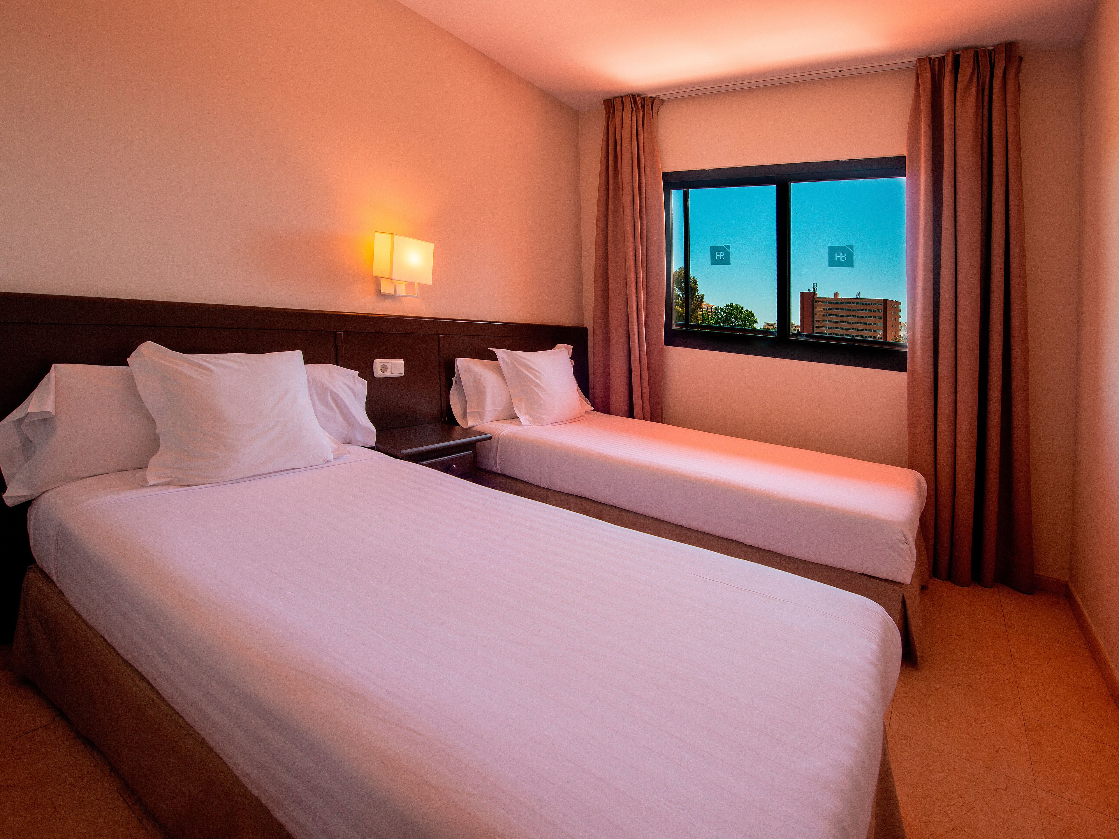 Habitación de hotel con dos camas, una mesita de noche, una lámpara y un ventanal con cortinas.