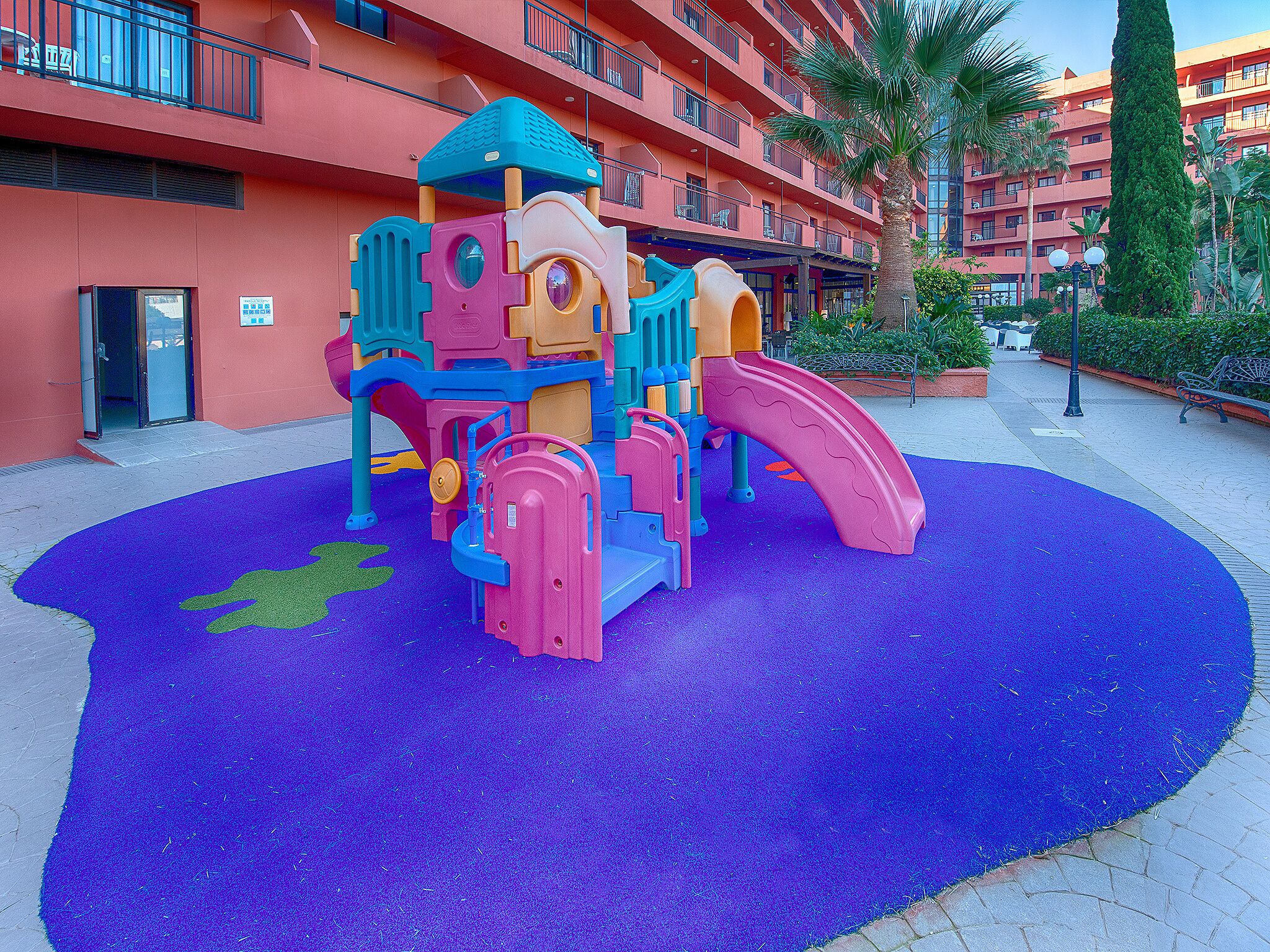 A playground with a slide and climbing structures, surrounded by a purple play area, in front of a multi-story building with balconies.