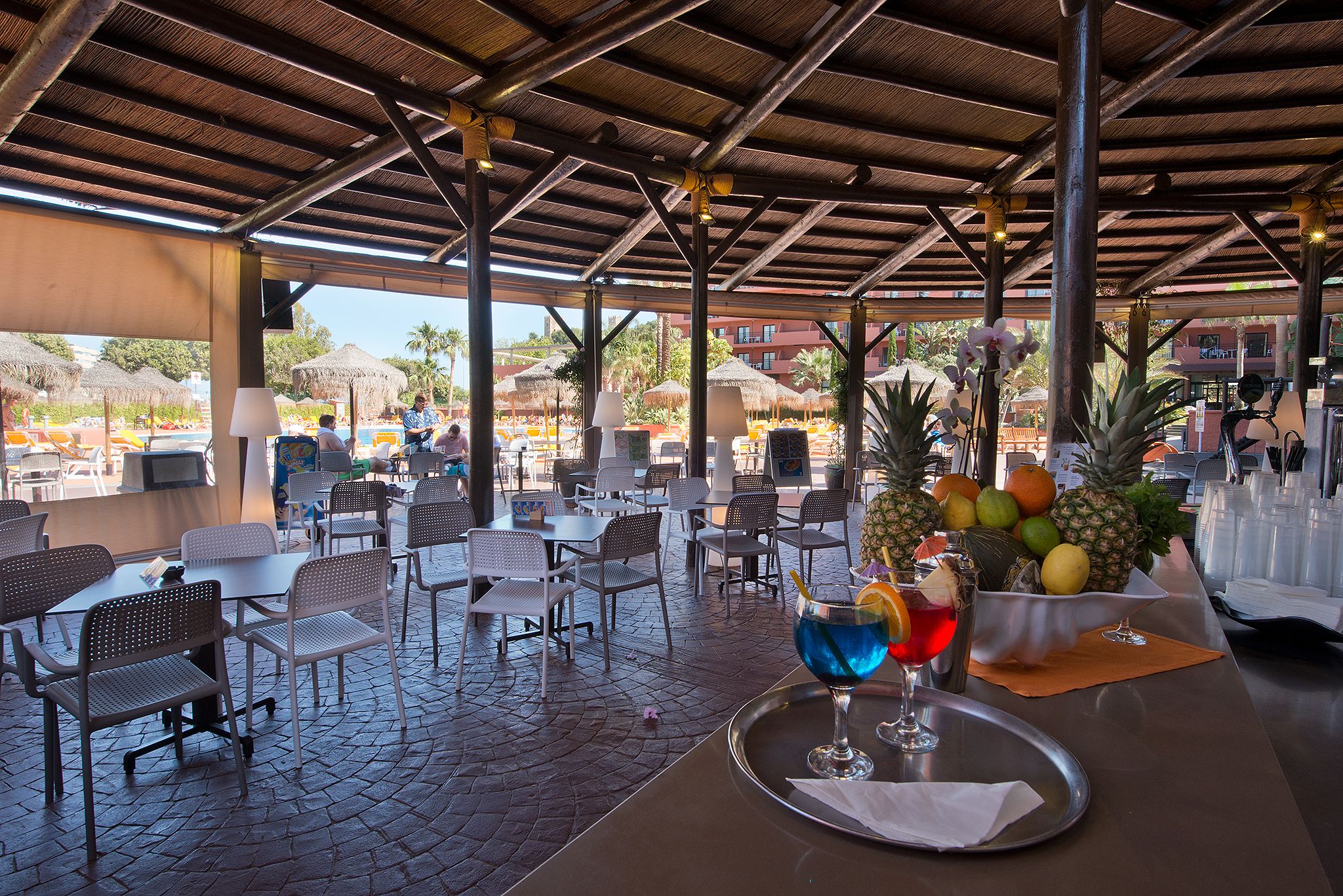 An outdoor dining area with tables and chairs, a tropical fruit display, and a drink on a tray.