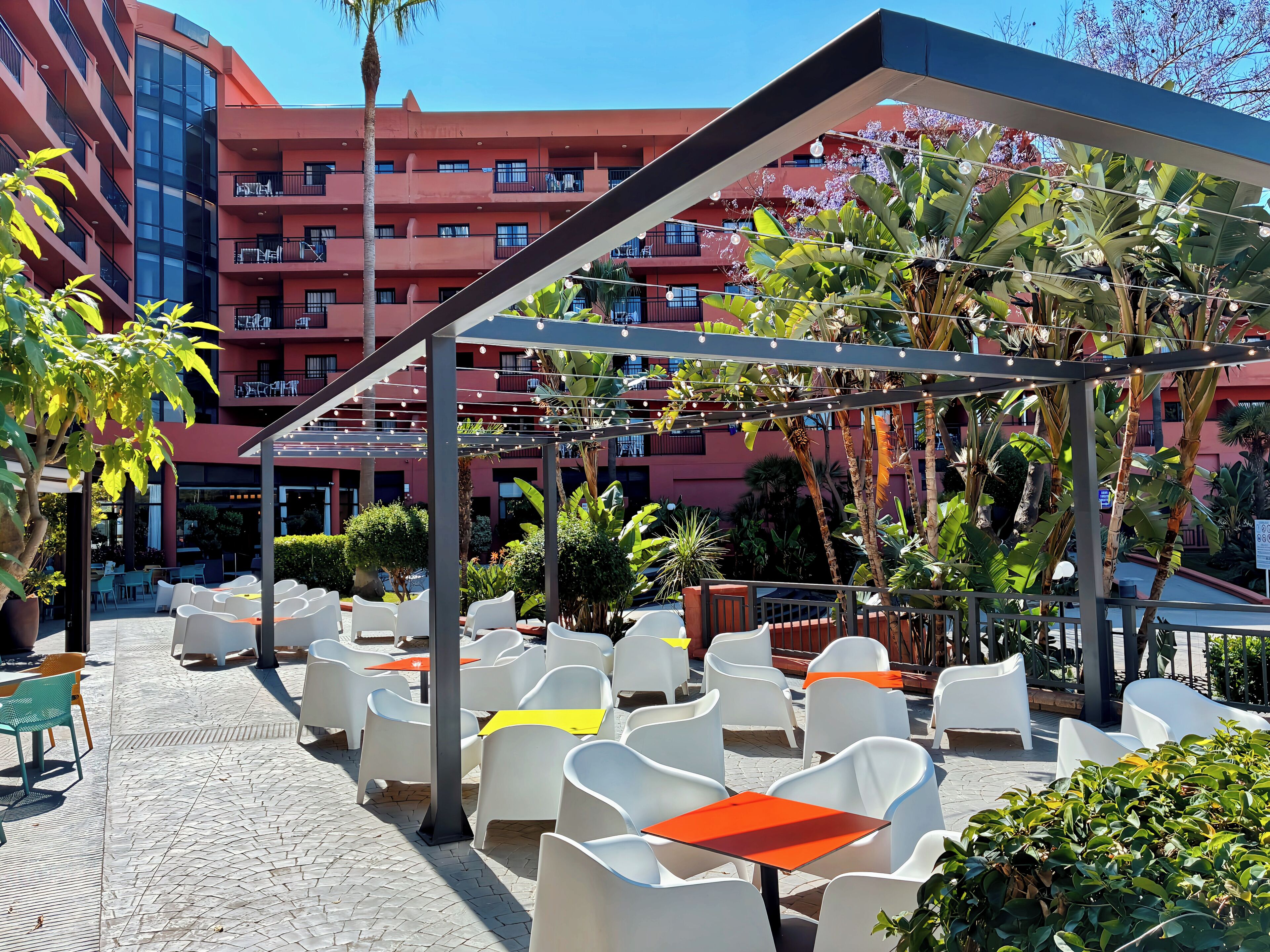 An outdoor patio area with white plastic chairs and tables, surrounded by lush greenery and a red building in the background.
