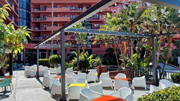 An outdoor patio area with white plastic chairs and tables, surrounded by lush greenery and a red building in the background.
