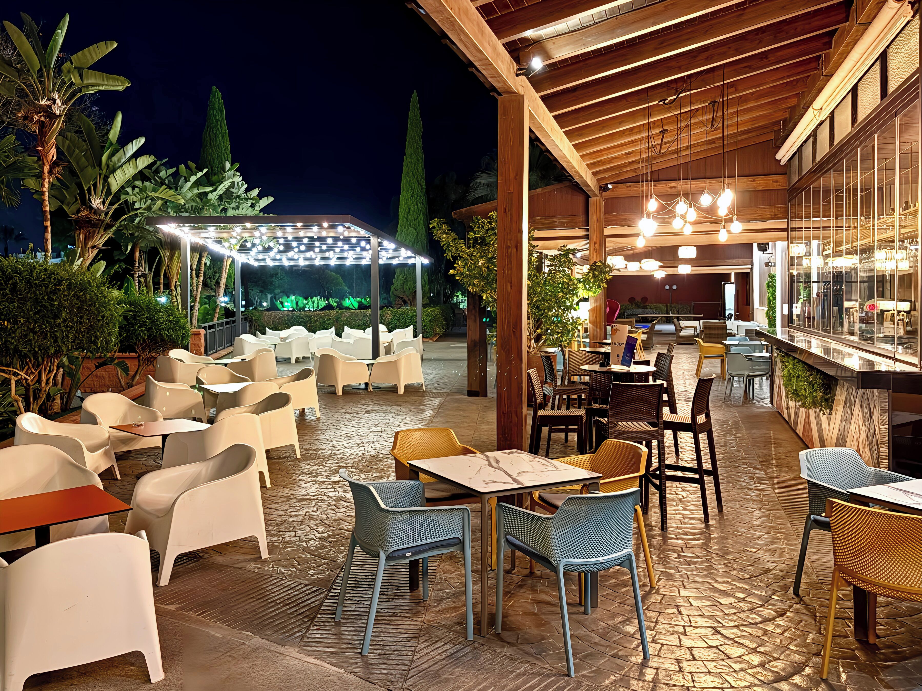 Un espace extérieur aménagé en terrasse, avec différents types de mobilier, notamment des tables et des chaises, placé sous une pergola en bois illuminée de guirlandes lumineuses.