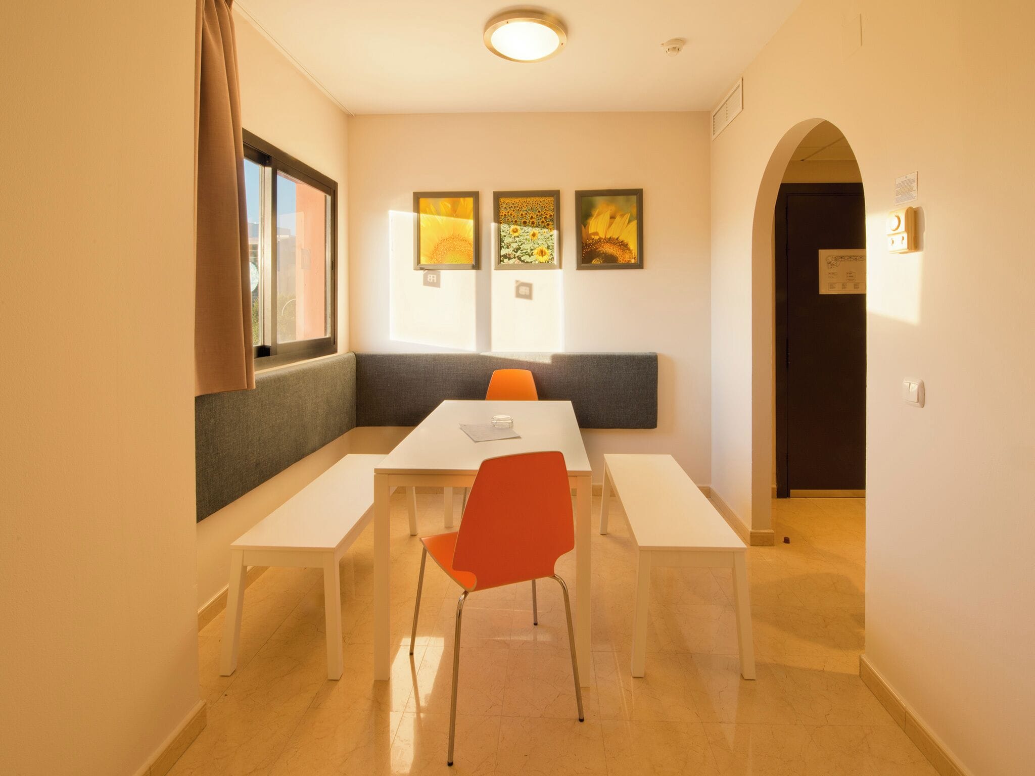 A modern dining area with a white table, orange chairs, and a bench. There are three framed artworks on the wall.