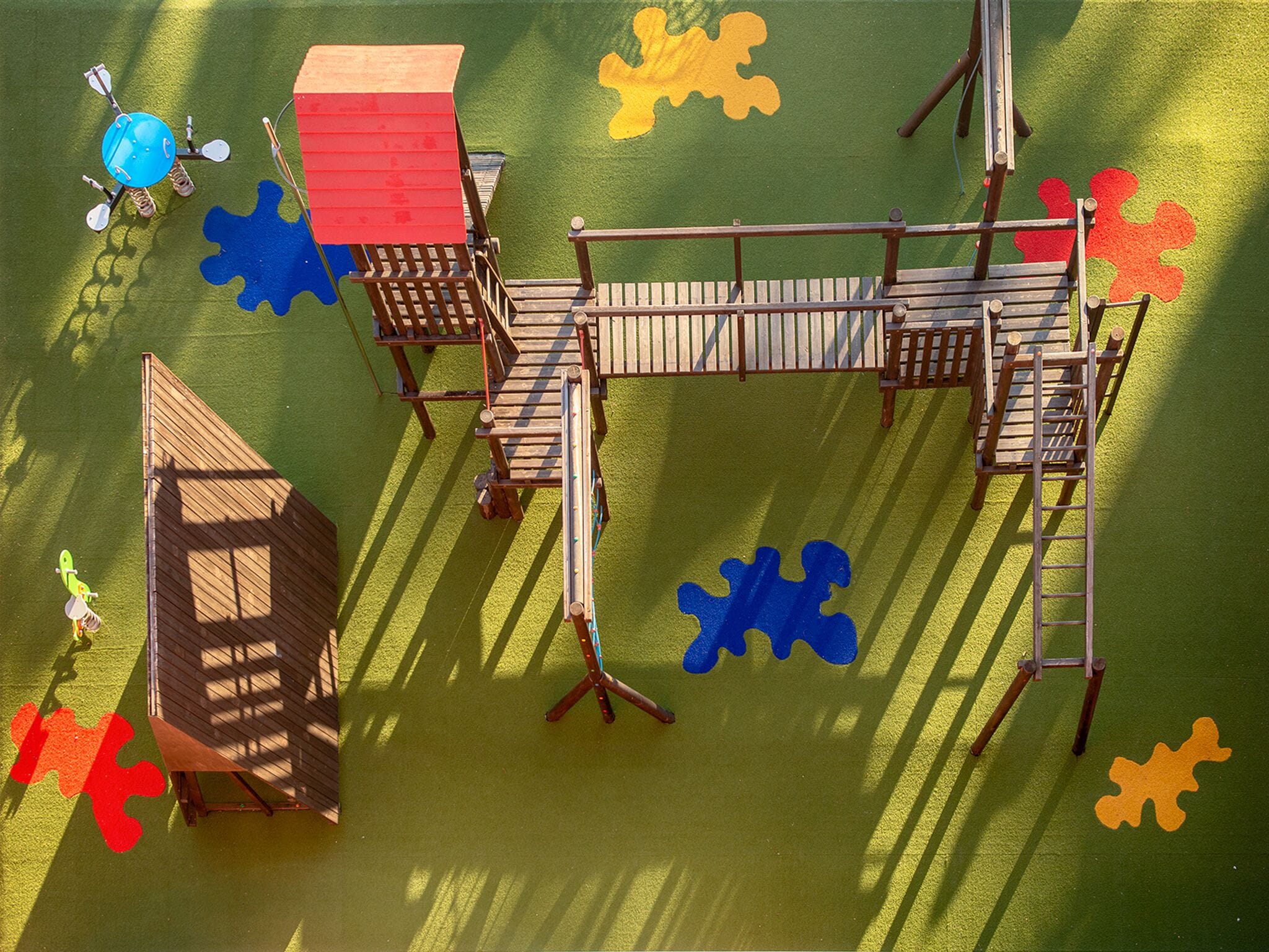 Une aire de jeux avec une structure d’escalade en bois, une maison de jeux au toit rouge et des marquages au sol en forme de pièces de puzzle.