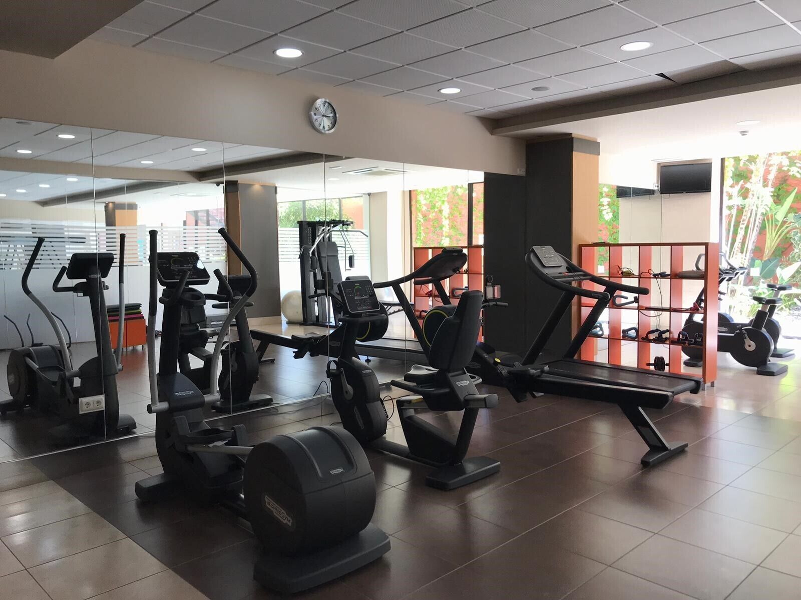 A gym with various exercise machines, a clock on the wall, and a view of greenery outside.