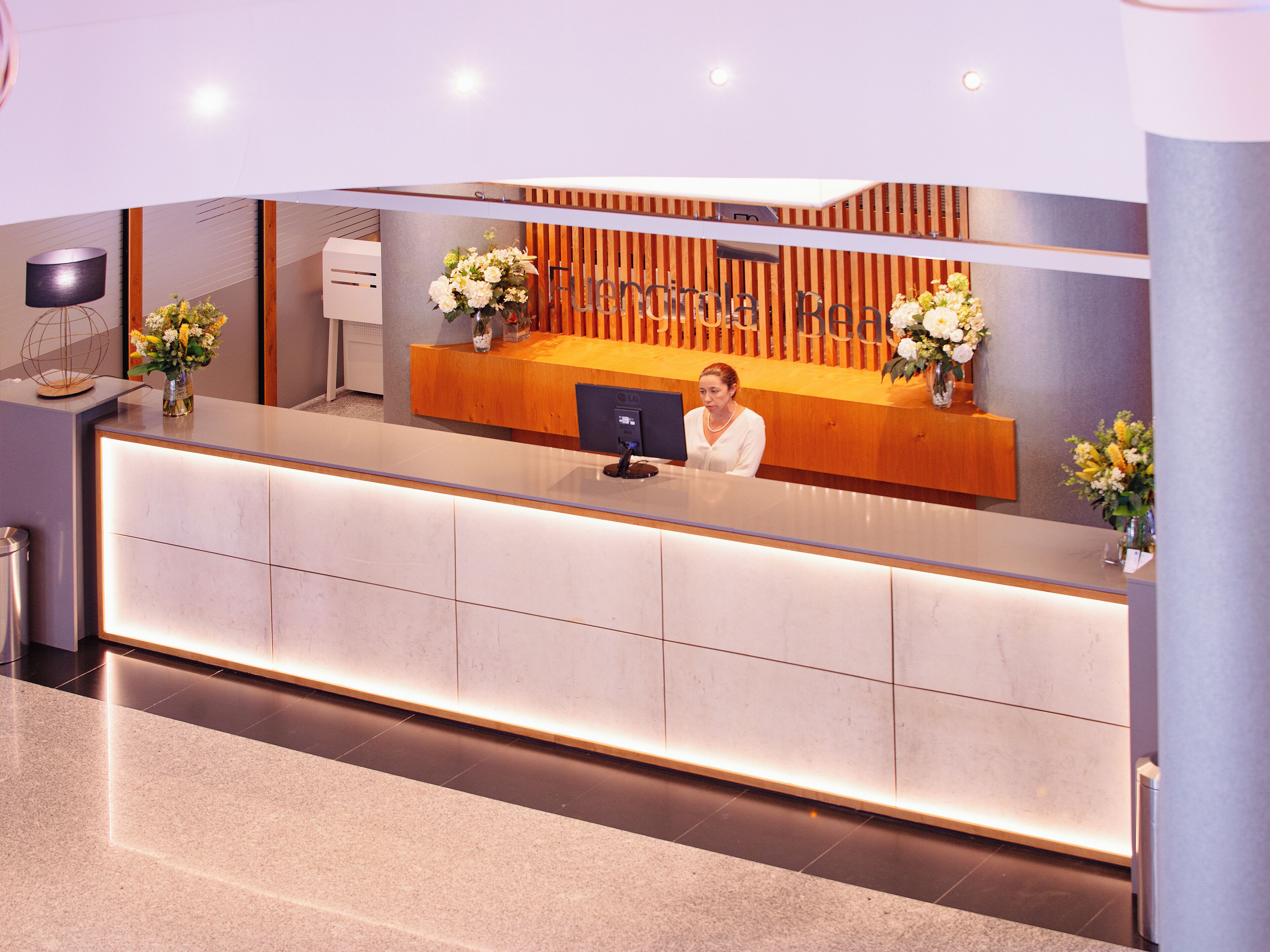 A reception desk with a computer, a wooden panel wall, and two flower arrangements.