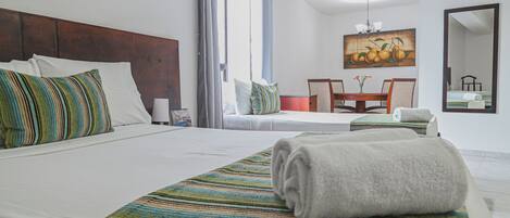 A hotel room with a large bed, a small table, and a chair. There is a window with curtains, a painting on the wall, and a mirror.