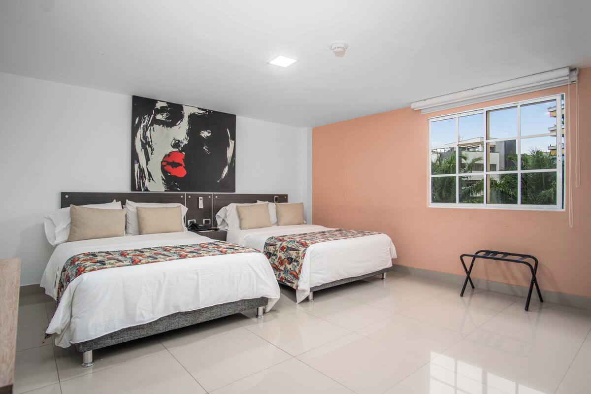 Quarto de hotel com duas camas, um quadro abstrato grande, uma janela com vista e uma mesa pequena.