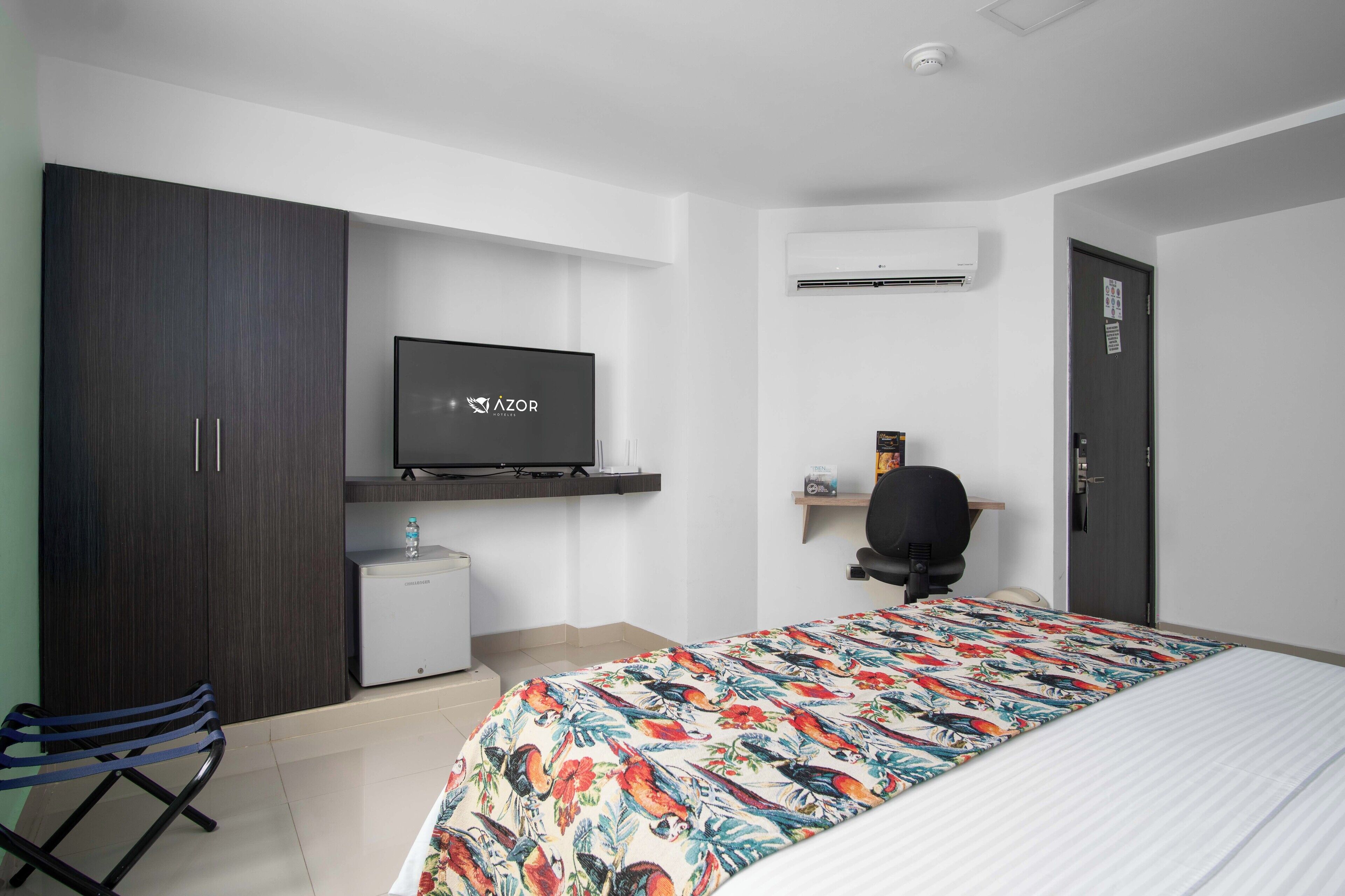 A hotel room with a bed, a TV, a desk, a chair, a wardrobe, and an air conditioning unit.