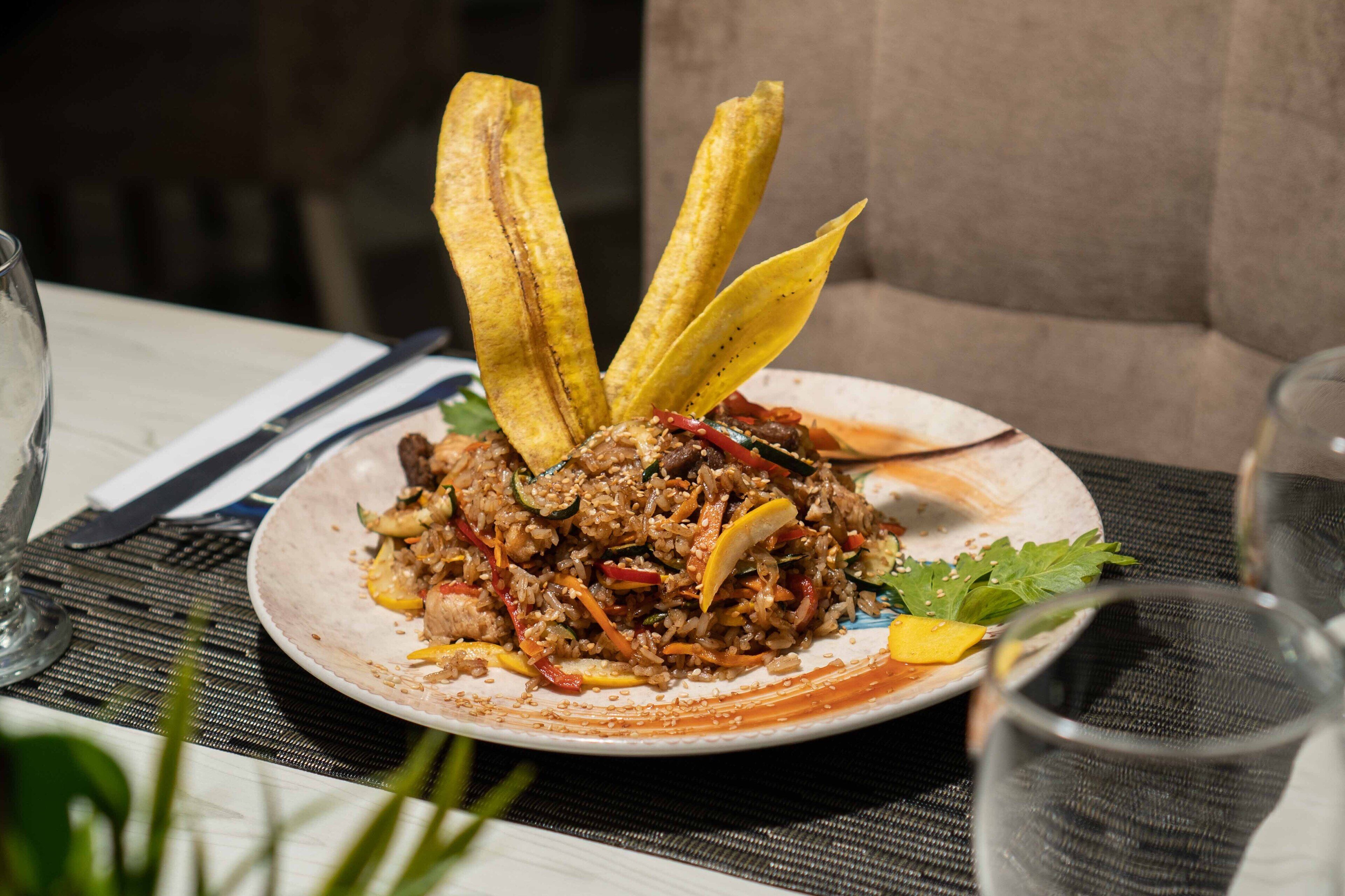 A plate of fried rice with vegetables and meat, accompanied by fried plantains, on a table with a placemat.