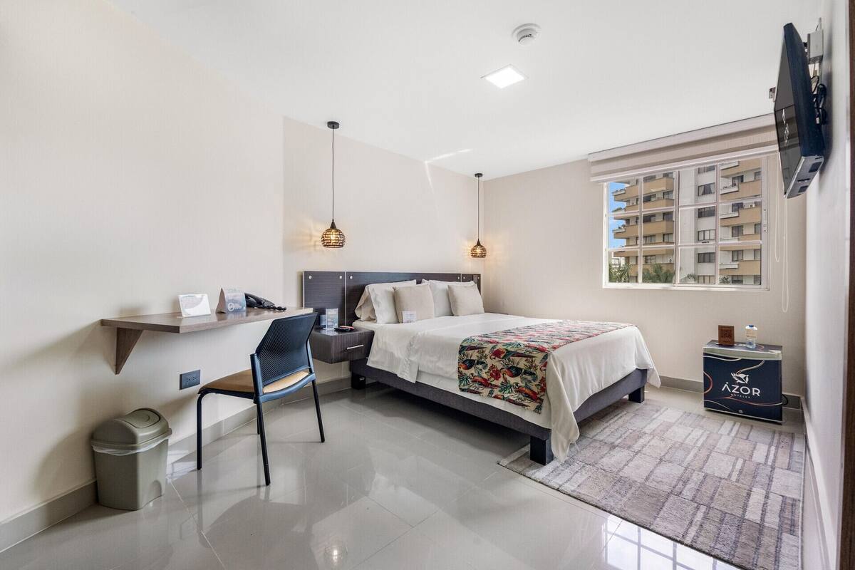 Quarto de hotel com cama, escrivaninha, cadeira, lixeira e TV fixada na parede.