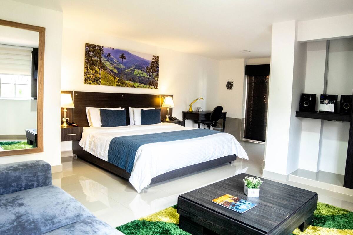 Quarto de hotel moderno com uma cama grande, uma escrivaninha, uma cadeira, uma mesa de centro e um quadro na parede.