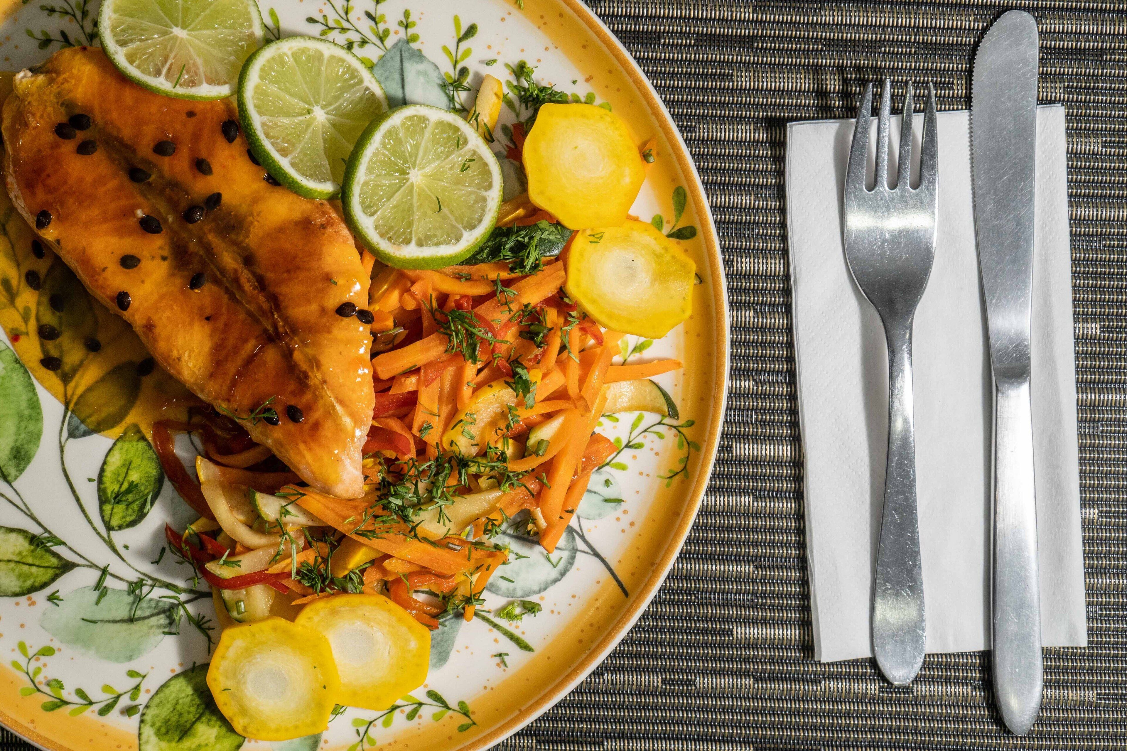 Un plato de comida con salmón, zanahorias y papas, junto a un tenedor y cuchillo sobre una mesa.