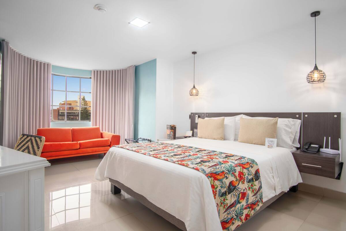 Quarto de hotel com cama grande, sofá, escrivaninha e janela com cortinas.