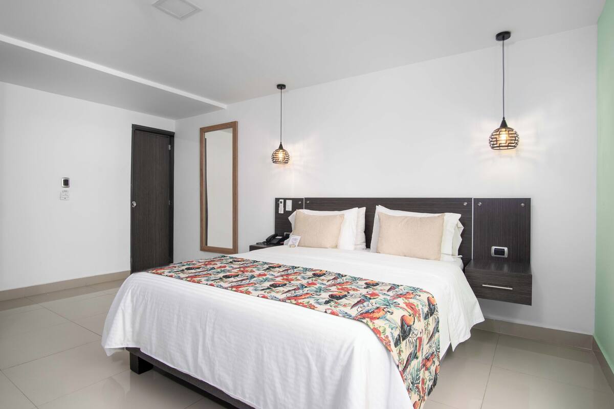 Uma cama bem arrumada com colcha estampada, uma mesinha de cabeceira com abajur e um painel de controle fixado na parede.