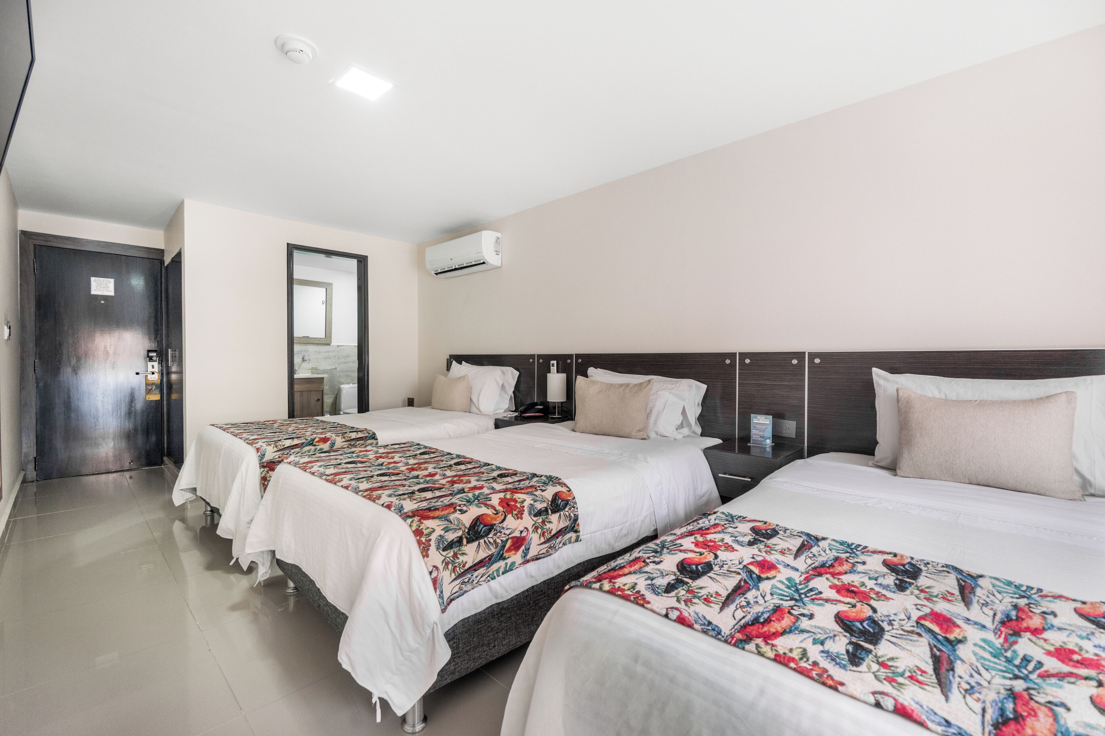 Habitación de hotel con tres camas, un cabecero de madera oscura, un equipo de aire acondicionado blanco y una puerta con picaporte.
