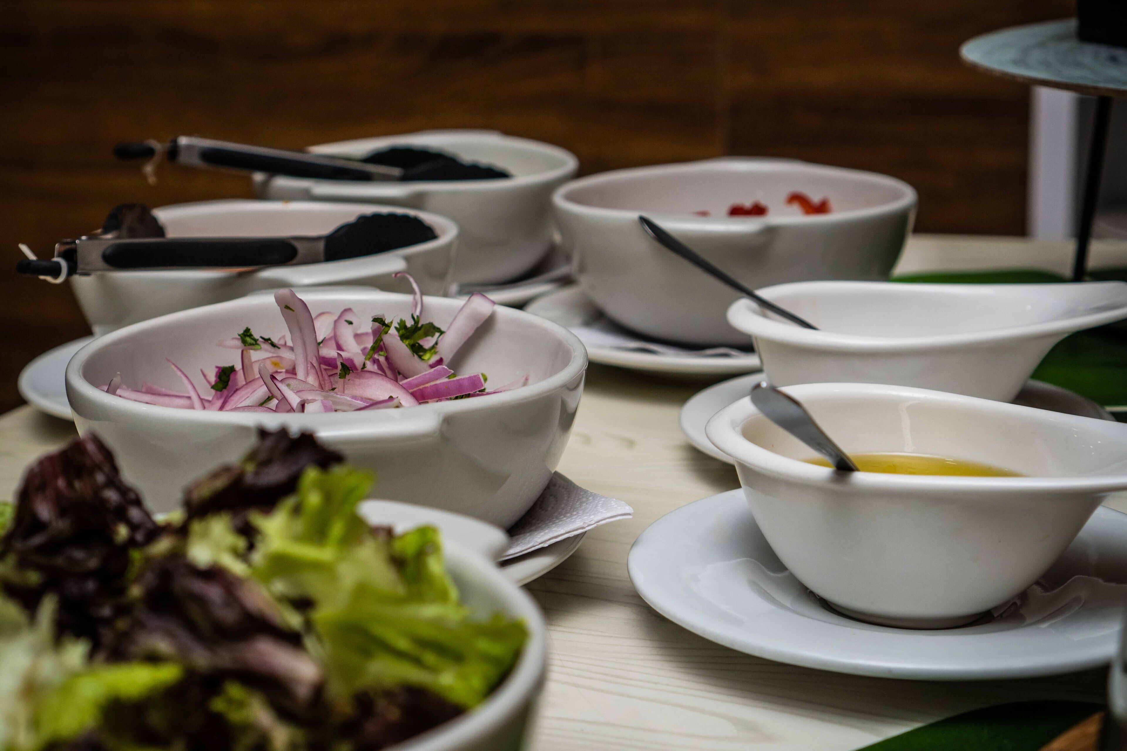 Ein Tisch mit verschiedenen Schalen, gefüllt mit Speisen, wobei einige Zutaten wie Zwiebeln und Salat sichtbar sind.