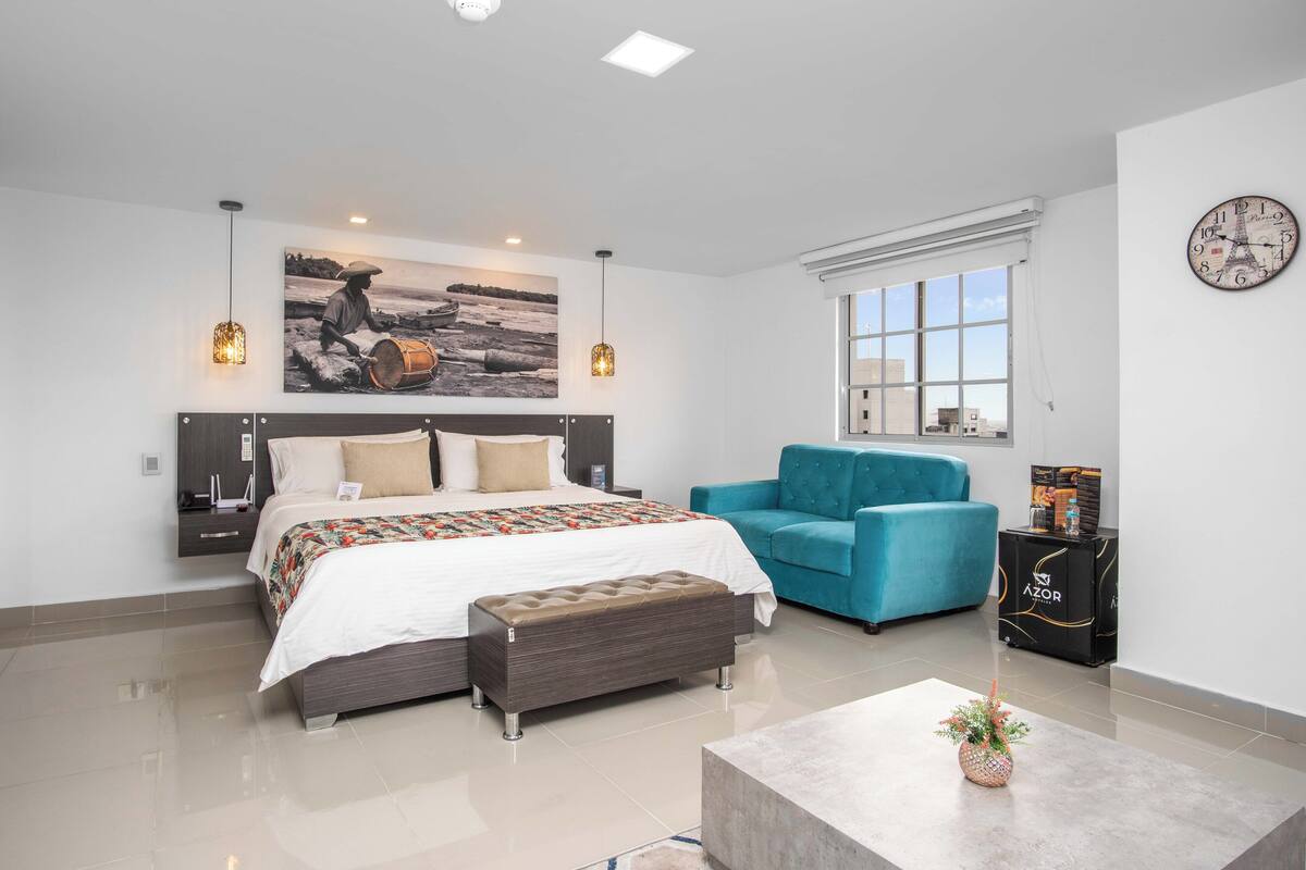 Quarto moderno com cama, banco, poltrona azul-esverdeada e mesa de centro.