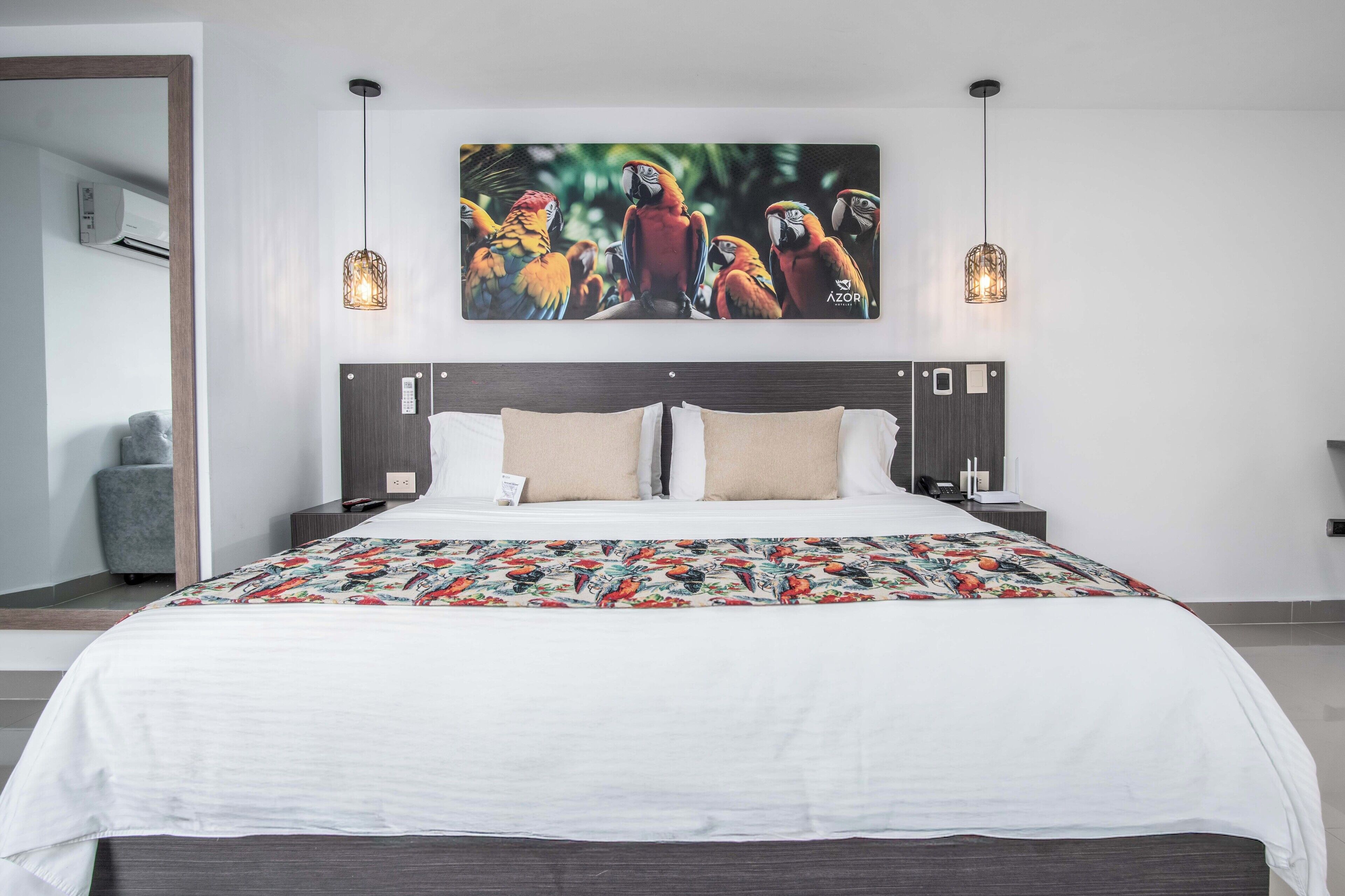 Una camera d'albergo moderna con un letto grande, una testiera con un pannello di controllo, due lampadari pendenti e un'opera d'arte colorata raffigurante pappagalli sulla parete.