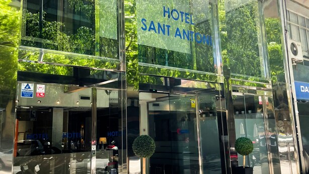Una entrada de hotel moderna con amplios ventanales de vidrio, un cartel que dice "HOTEL SANT ANTONI" y una puerta de acceso visible.