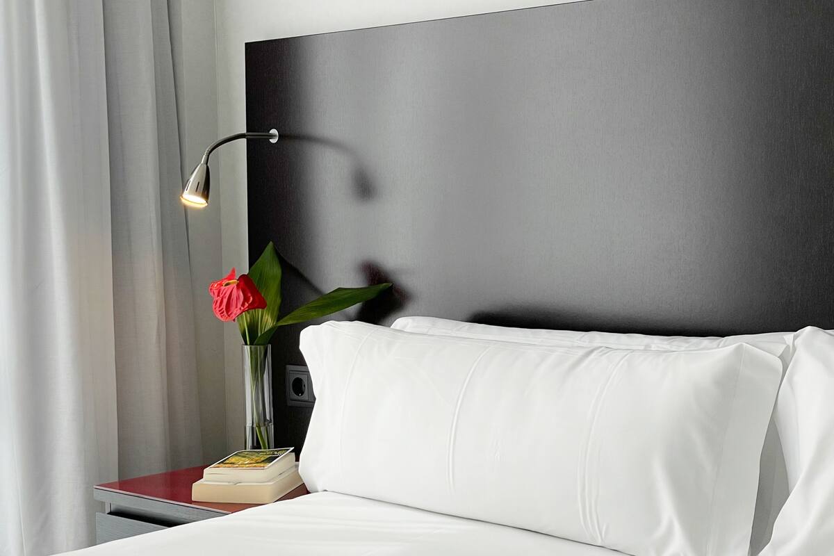 Ein ordentlich bezogenes Bett mit weißen Leinen, ein Nachttisch mit Buch und rotem Gegenstand, eine Vase mit roter Blume und eine moderne Wand mit dunklem Panel.