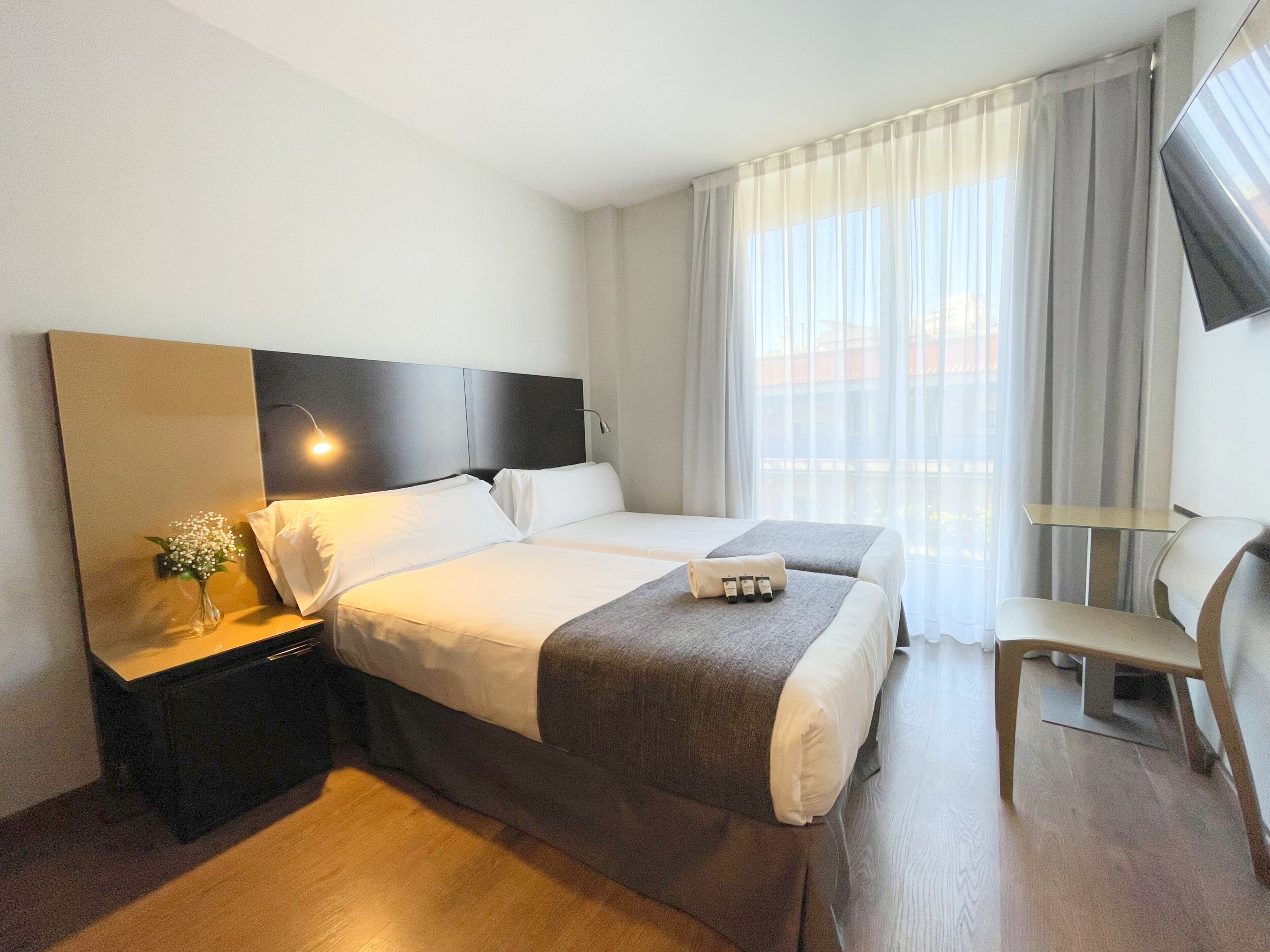 A hotel room with two beds, a bedside table with a lamp, a chair, and a TV on the wall.