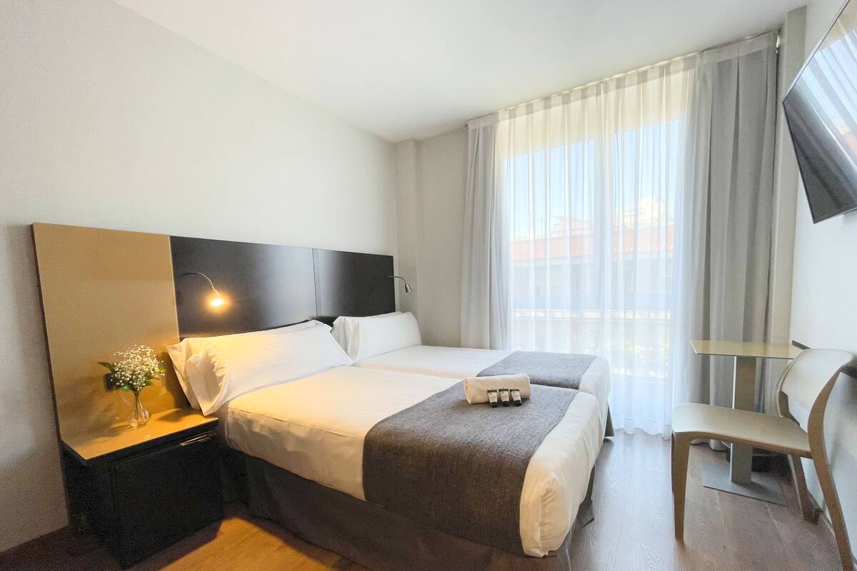 Ein Hotelzimmer mit zwei Betten, einem Nachttisch mit Lampe, einem Sessel und einem an der Wand befestigten Fernseher.