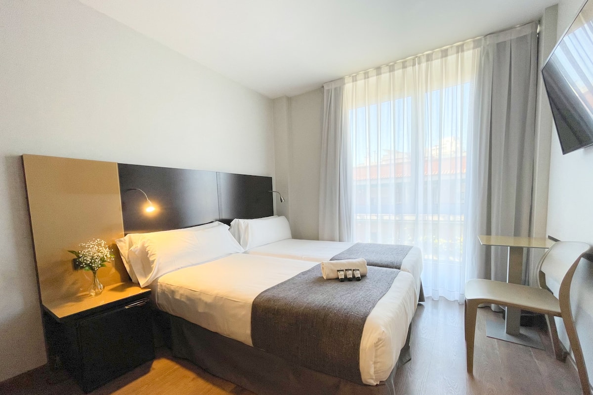 Habitación de hotel con dos camas, una mesita de noche con lámpara, una silla y un televisor en la pared.