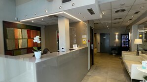 Un área de recepción moderna con un separador de vidrio, un mostrador de recepción, una orquídea en maceta y un rincón de espera con una lámpara.