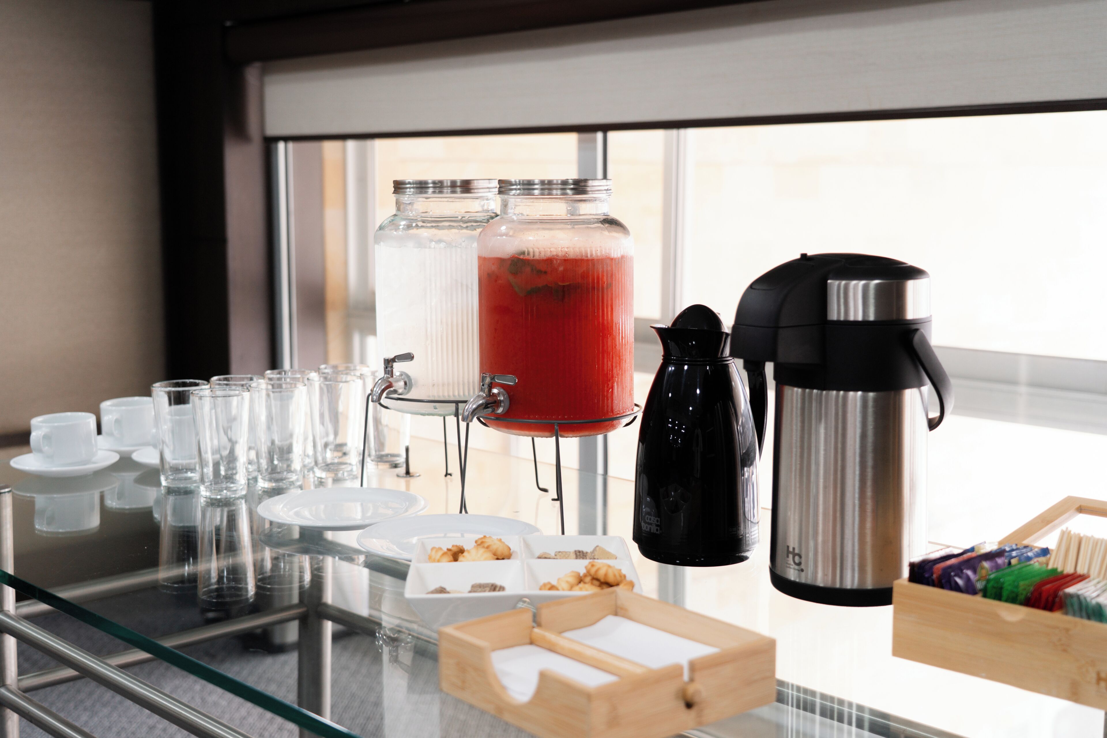 Una mesa de vidrio con una cafetera, un termo y un dispensador de bebida de frutas.